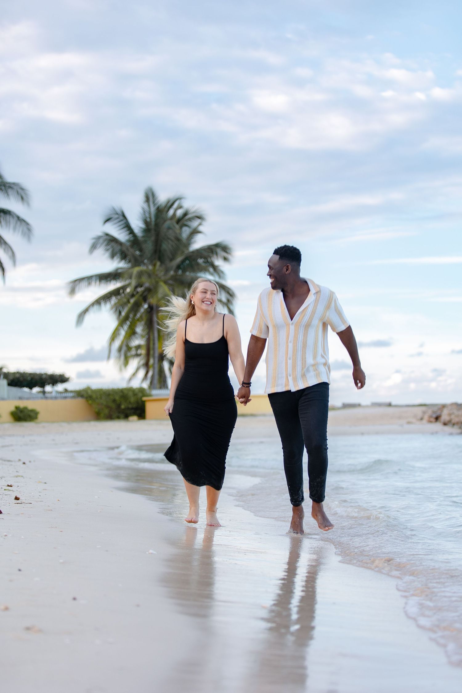 Joyful interracial couple walking barefoot along tropical beach with palm trees and cloudy sky.