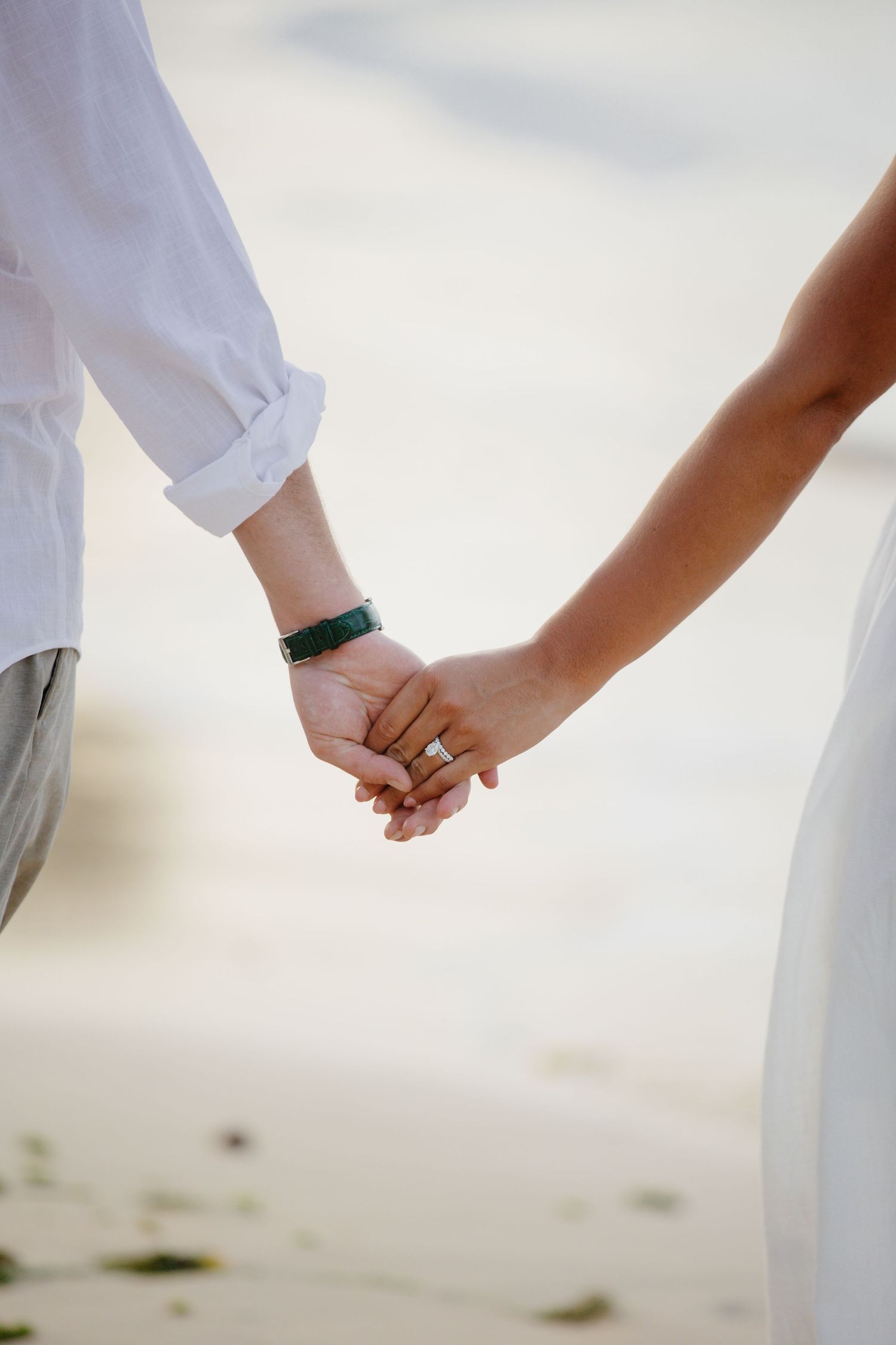 Couple holding hands on beach, woman wearing diamond engagement ring, both dressed in white, romantic wedding photo.