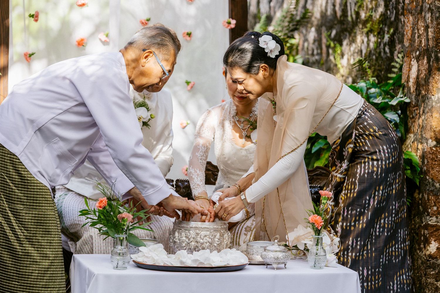 Elderly couple and wedding guest cutting cake together at an outdoor wedding reception under trees.