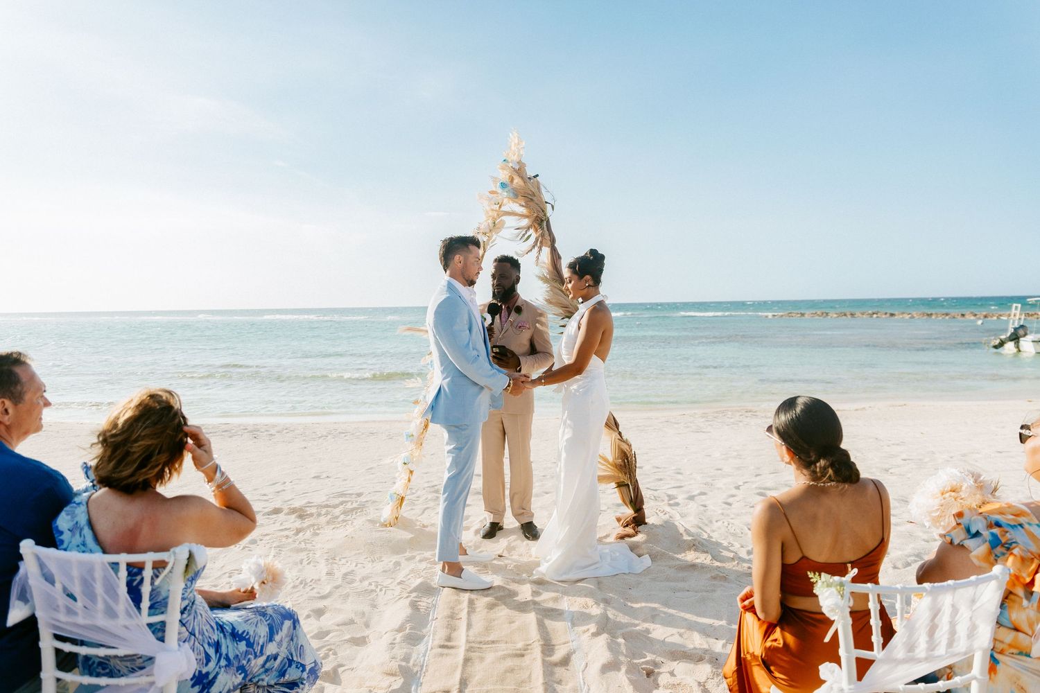 Interracial couple exchanging vows at beach wedding ceremony with boho pampas arch and ocean backdrop.