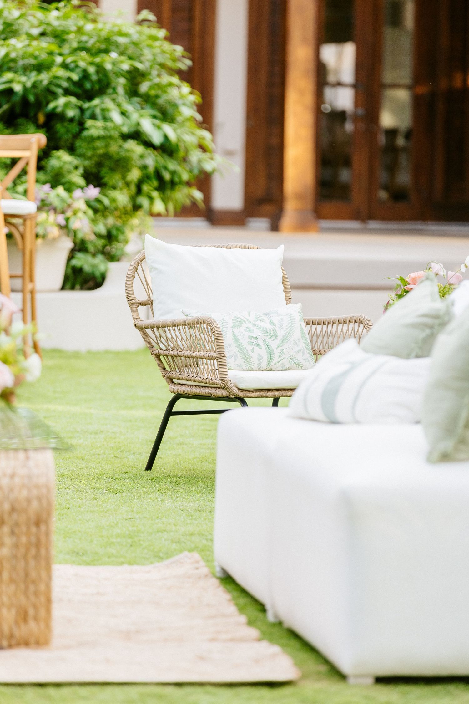 Elegant outdoor patio seating area with wicker chair and white sofa, green cushions on lush lawn near wooden doors.