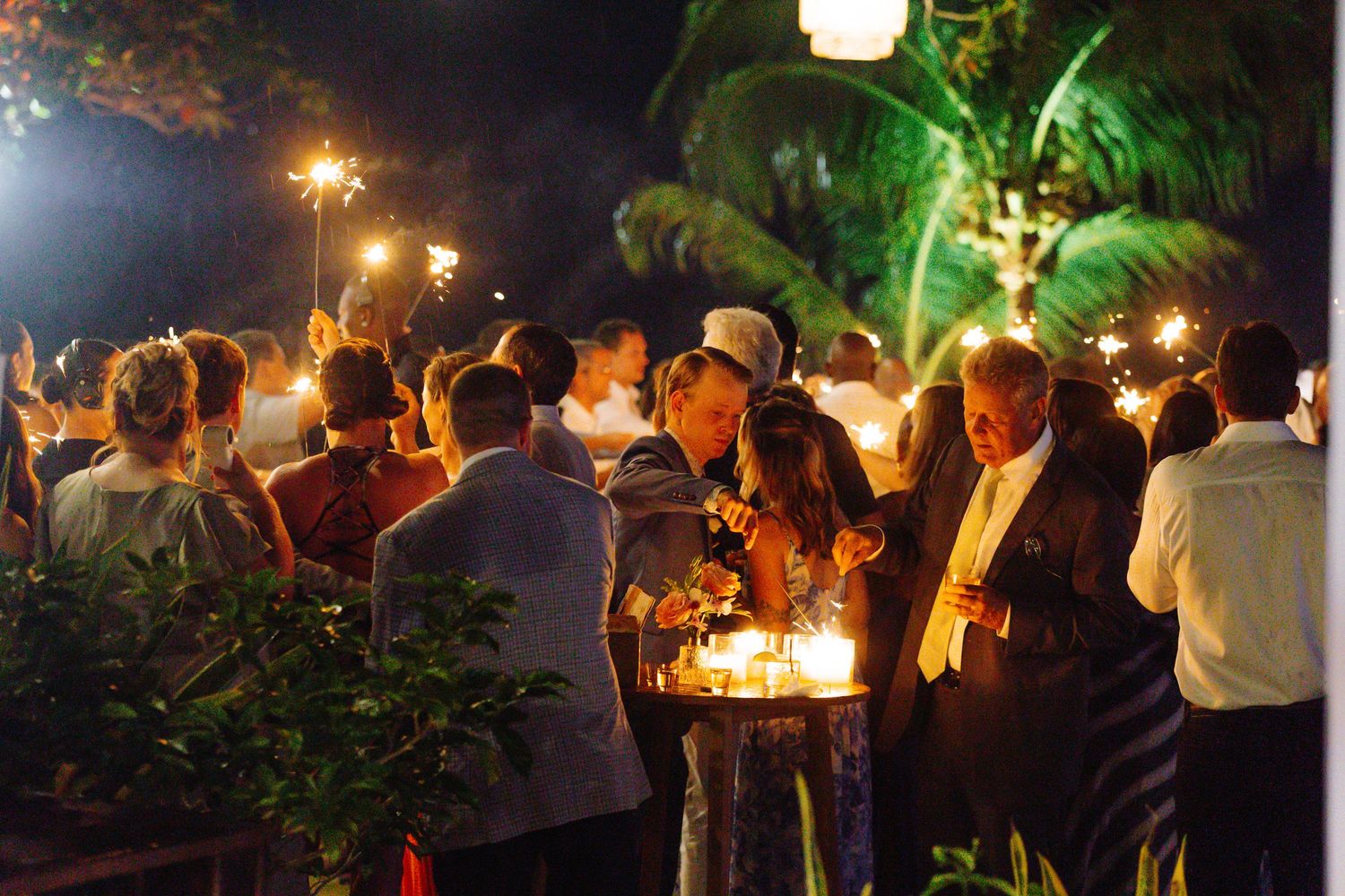 Elegant outdoor evening party with guests holding sparklers under palm trees at a festive night celebration.