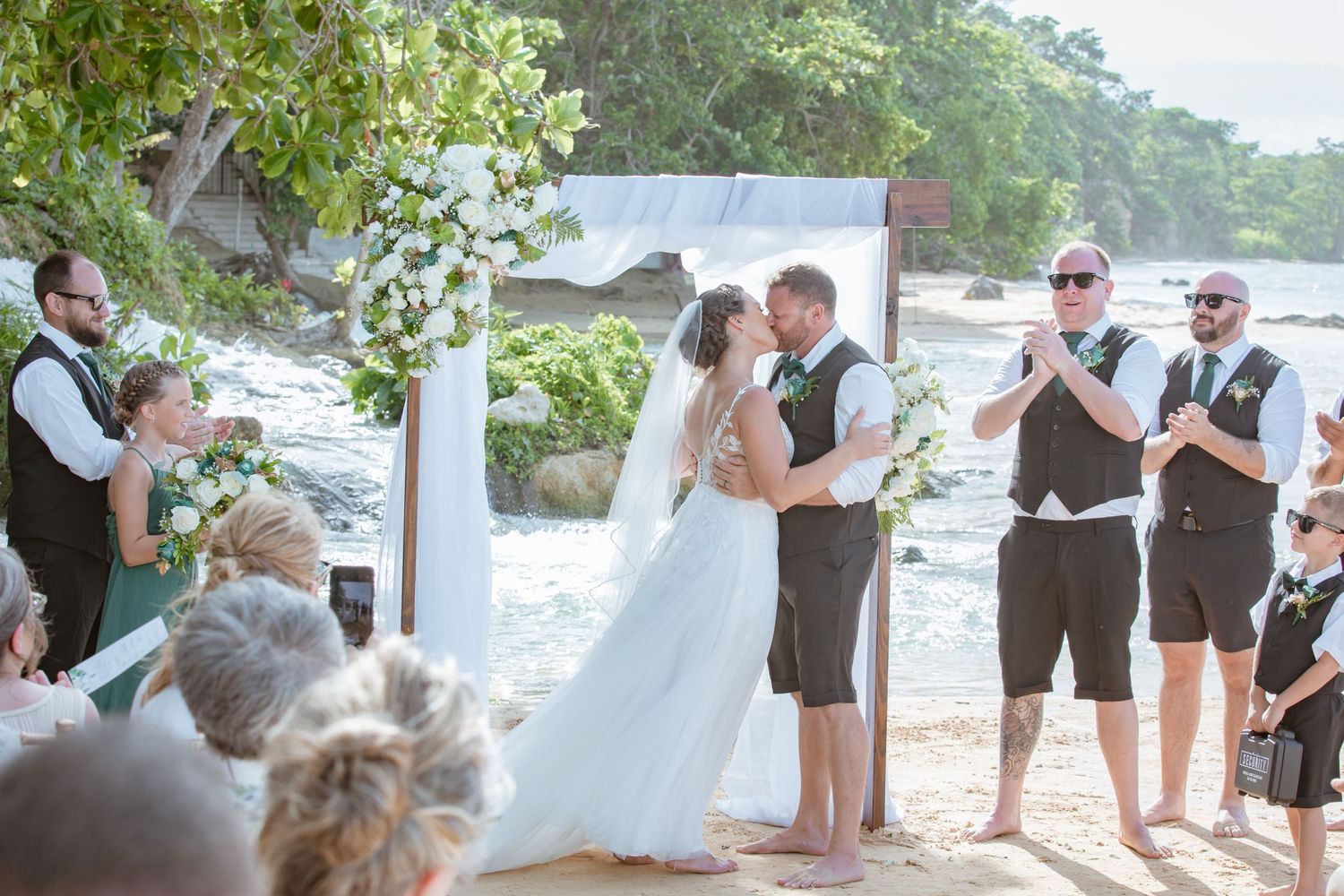 Bride and groom share first kiss at beach wedding altar surrounded by groomsmen and guests.