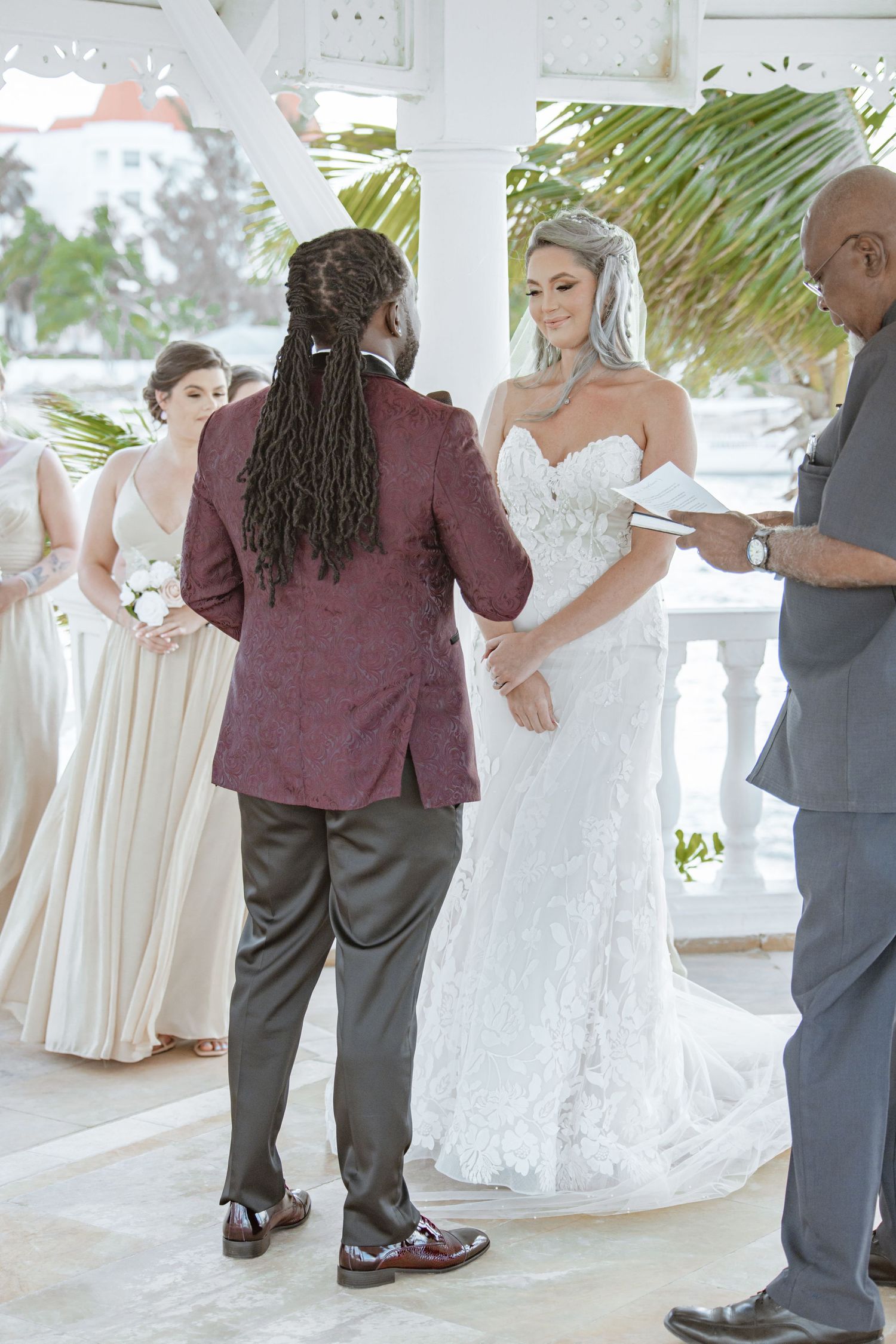 Groom in burgundy jacket exchanges vows with bride in white lace dress at elegant outdoor ceremony.