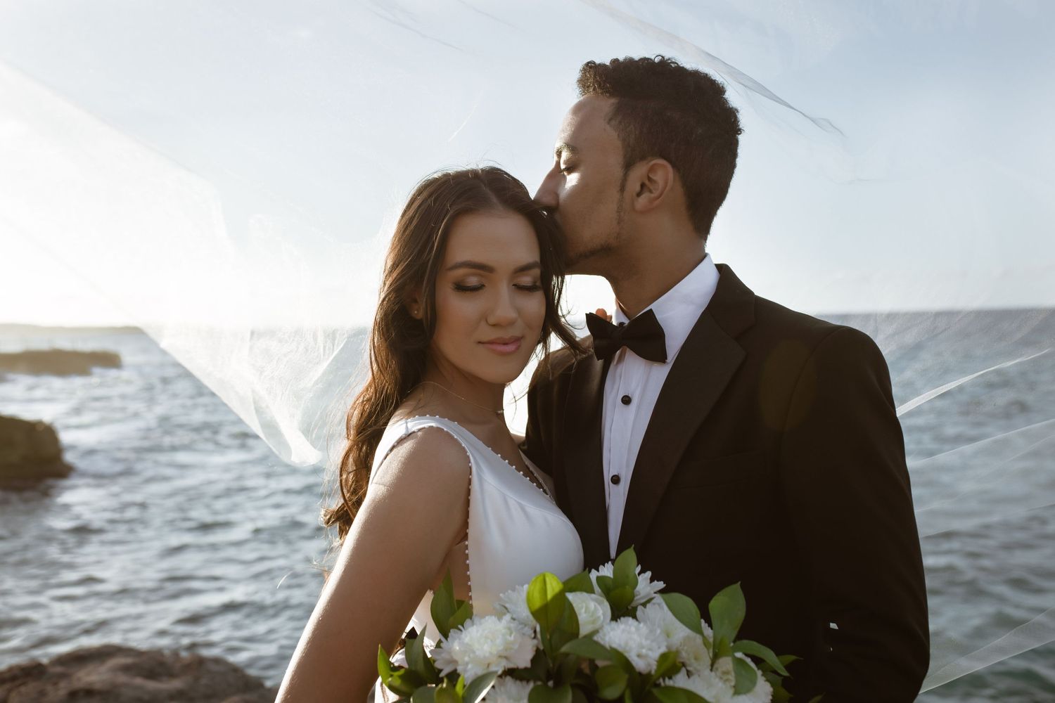 Bride and groom sharing romantic kiss by ocean at sunset, bride holding white bouquet.