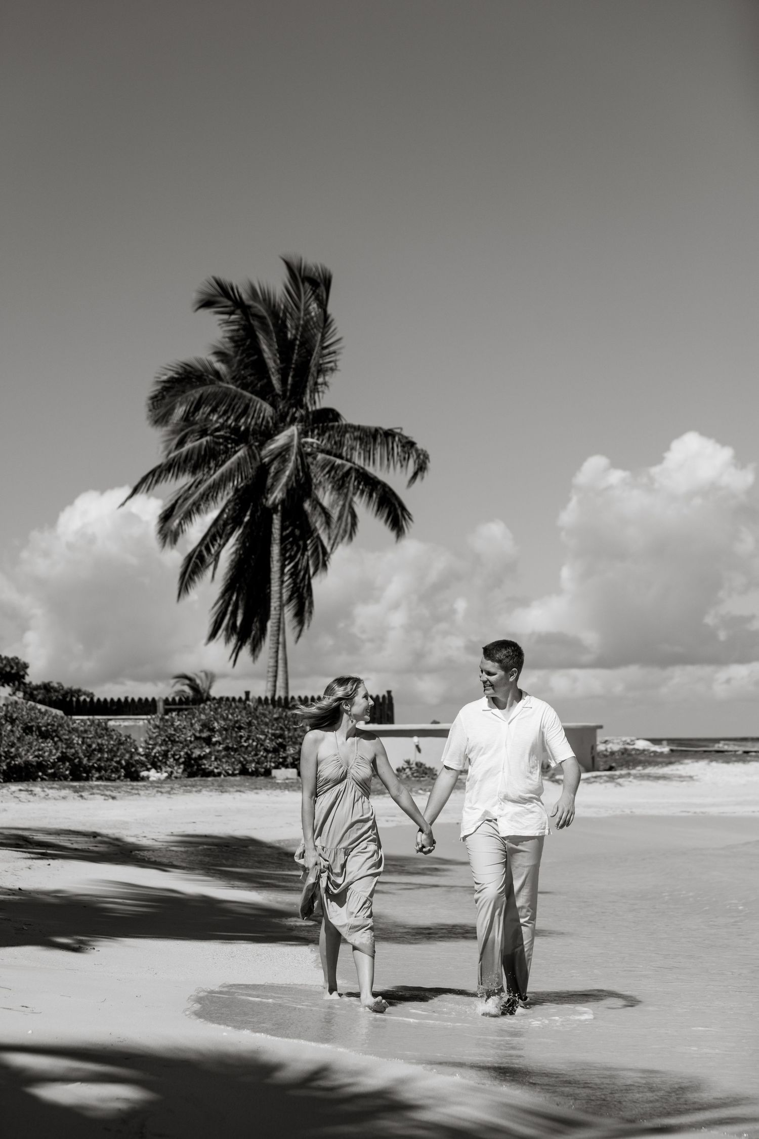 Black and white photo of couple holding hands walking on tropical beach near palm tree.
