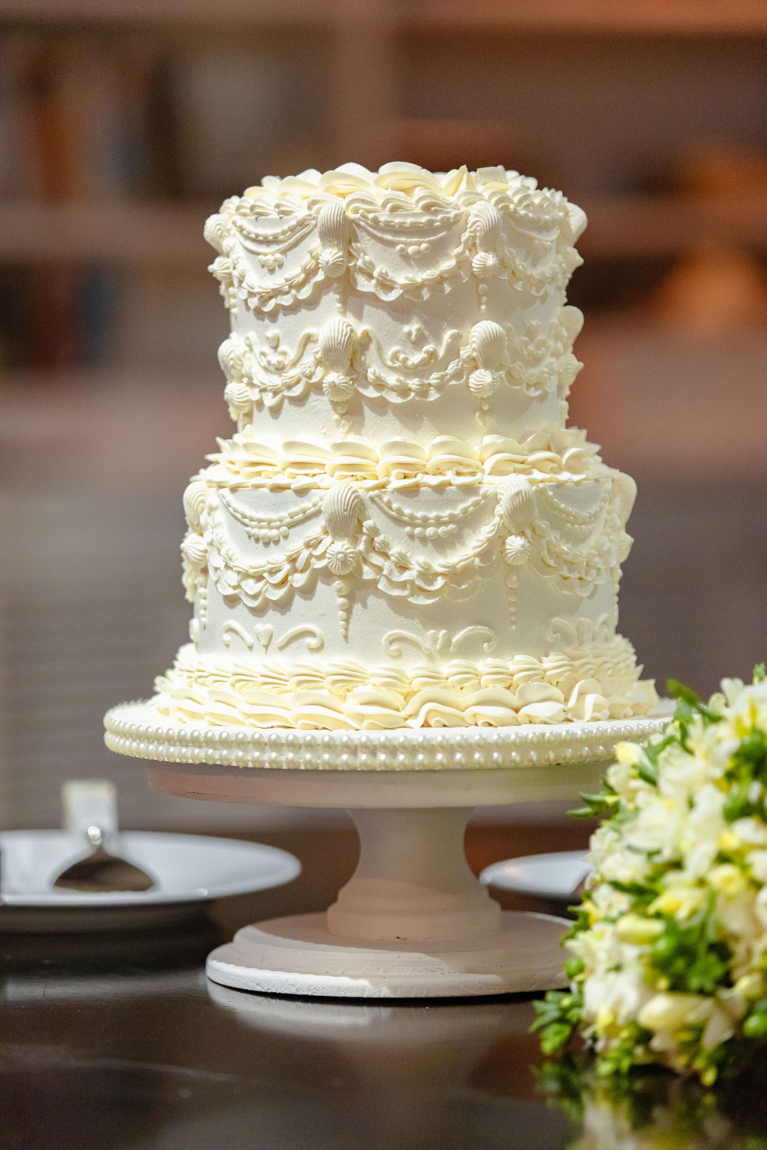 Two-tier white wedding cake with intricate pearl and swag piped frosting details on a pedestal.
