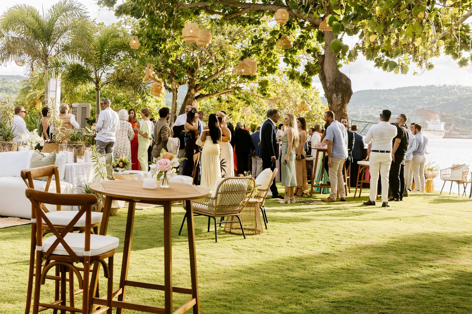 Outdoor wedding reception on lush green lawn with guests mingling under trees near a waterfront venue.