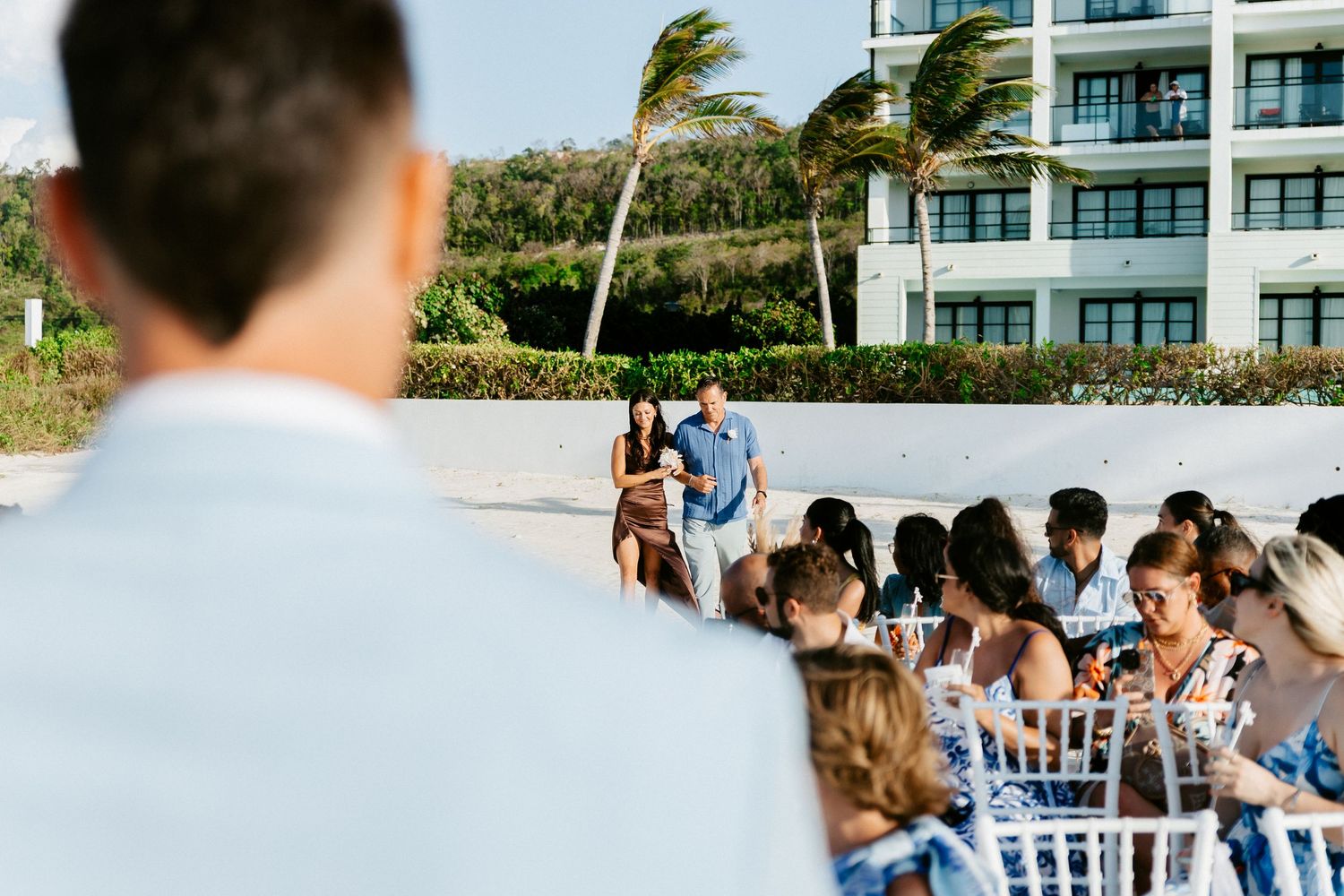 Outdoor beach resort wedding ceremony with guests seated in white chairs, palm trees swaying in the wind near a modern hotel.
