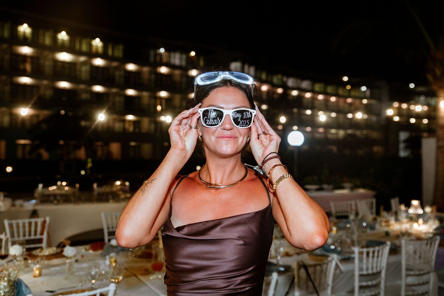 Smiling wedding guest in brown satin dress posing with white sunglasses at night reception.