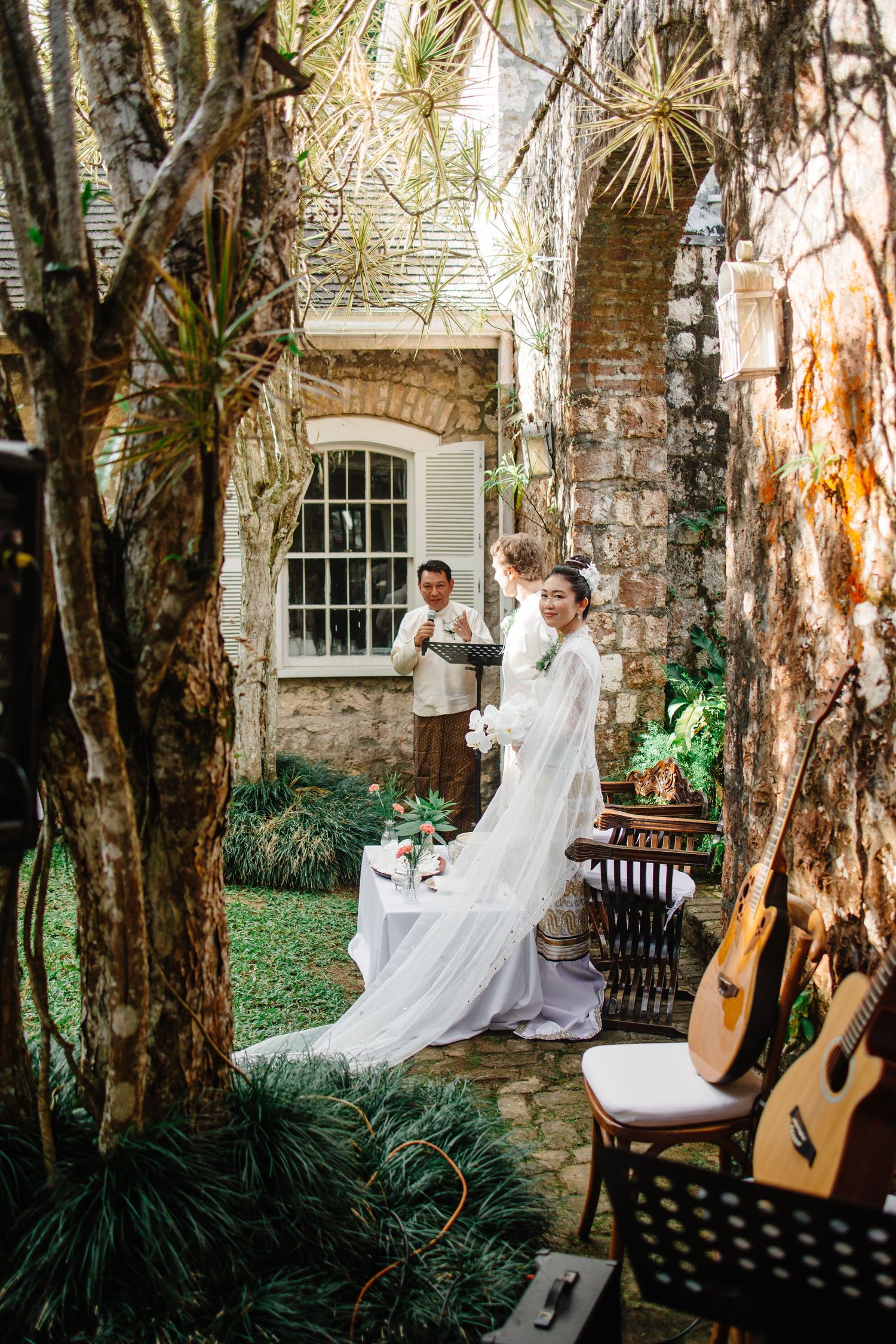 Intimate garden wedding ceremony with bride in white gown, officiant, and acoustic guitars among stone walls.