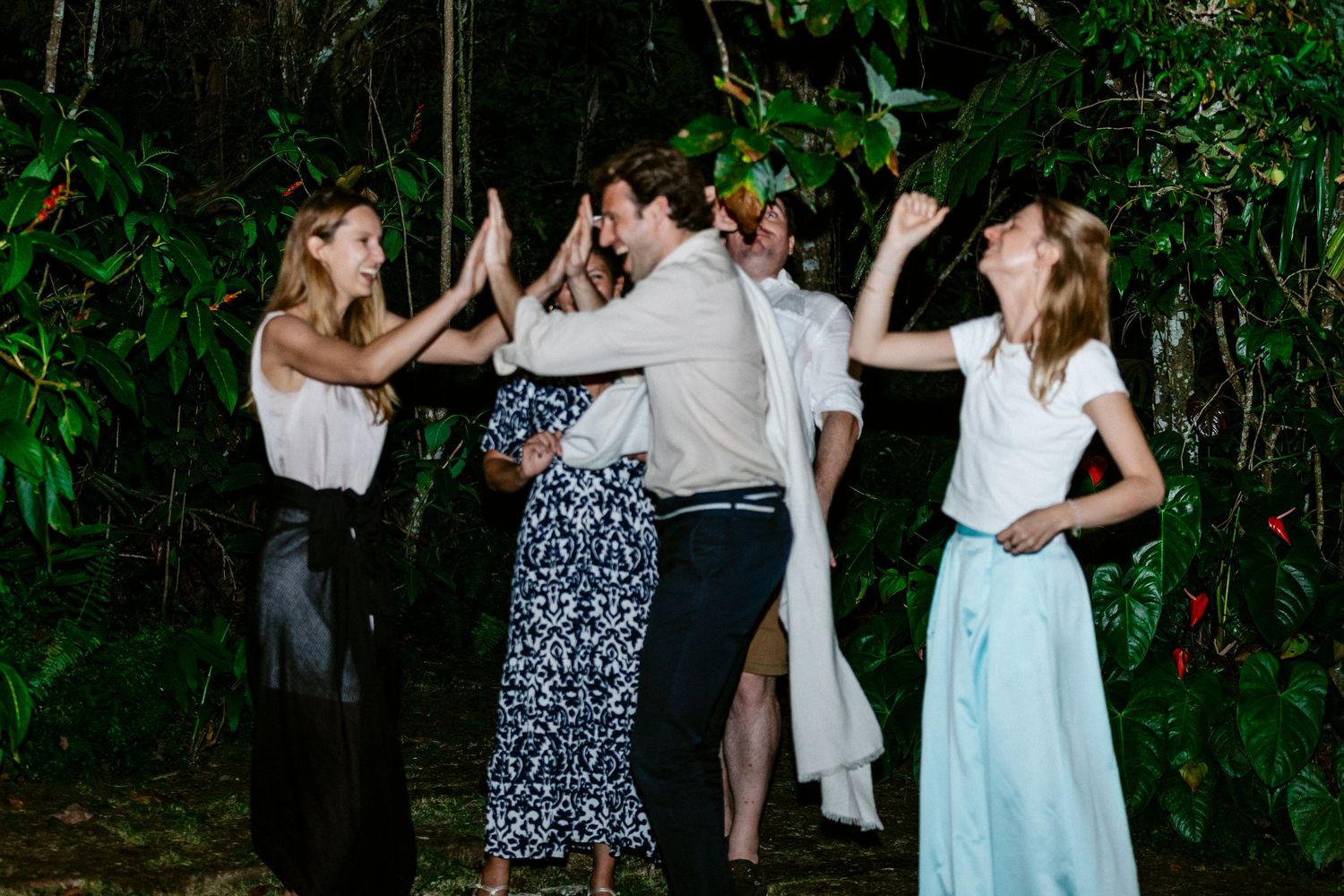 Group of friends dancing and celebrating outdoors at a tropical night party, wearing elegant attire.