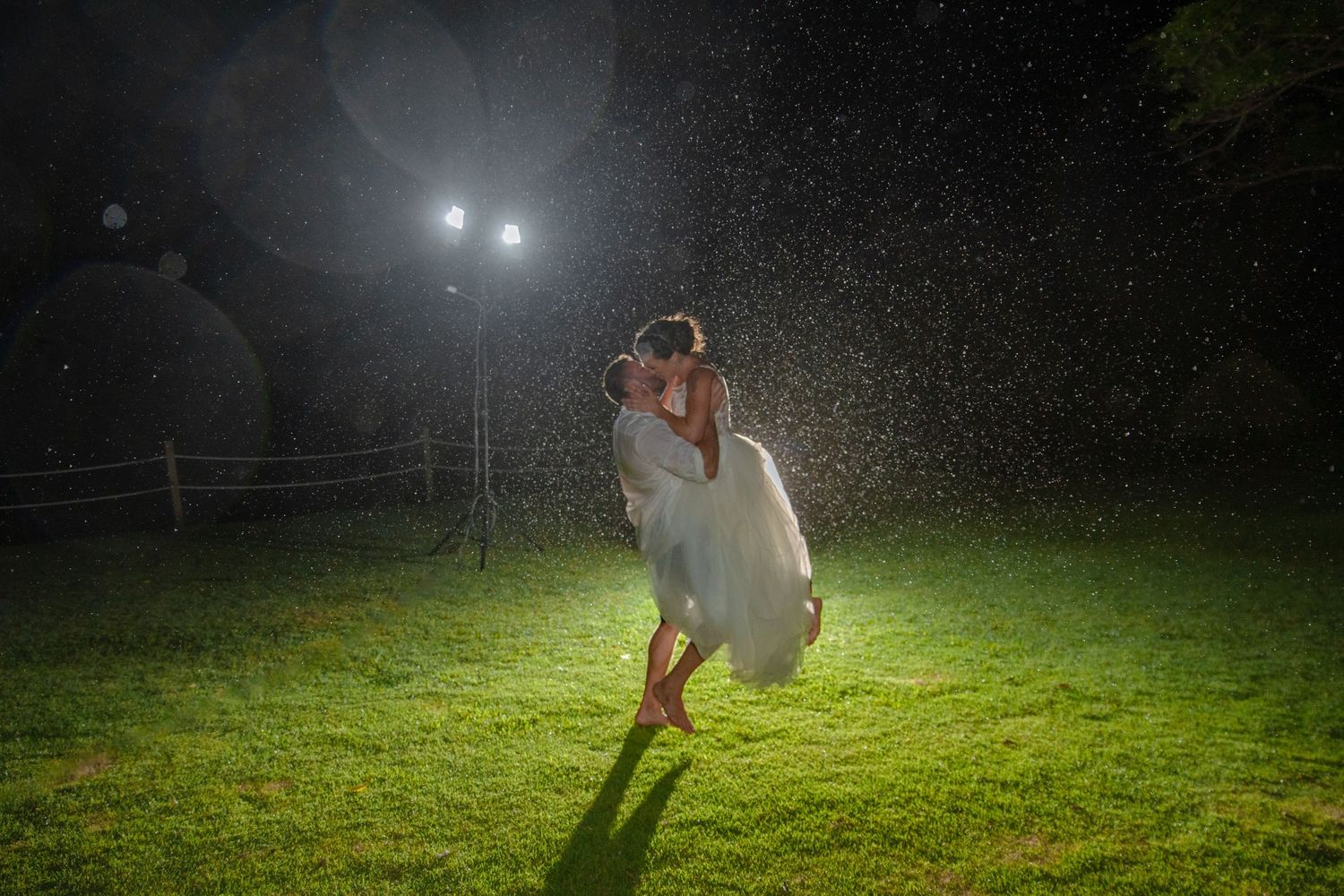 Couple sharing a romantic kiss in the rain at night, illuminated by bright lights on a green lawn.