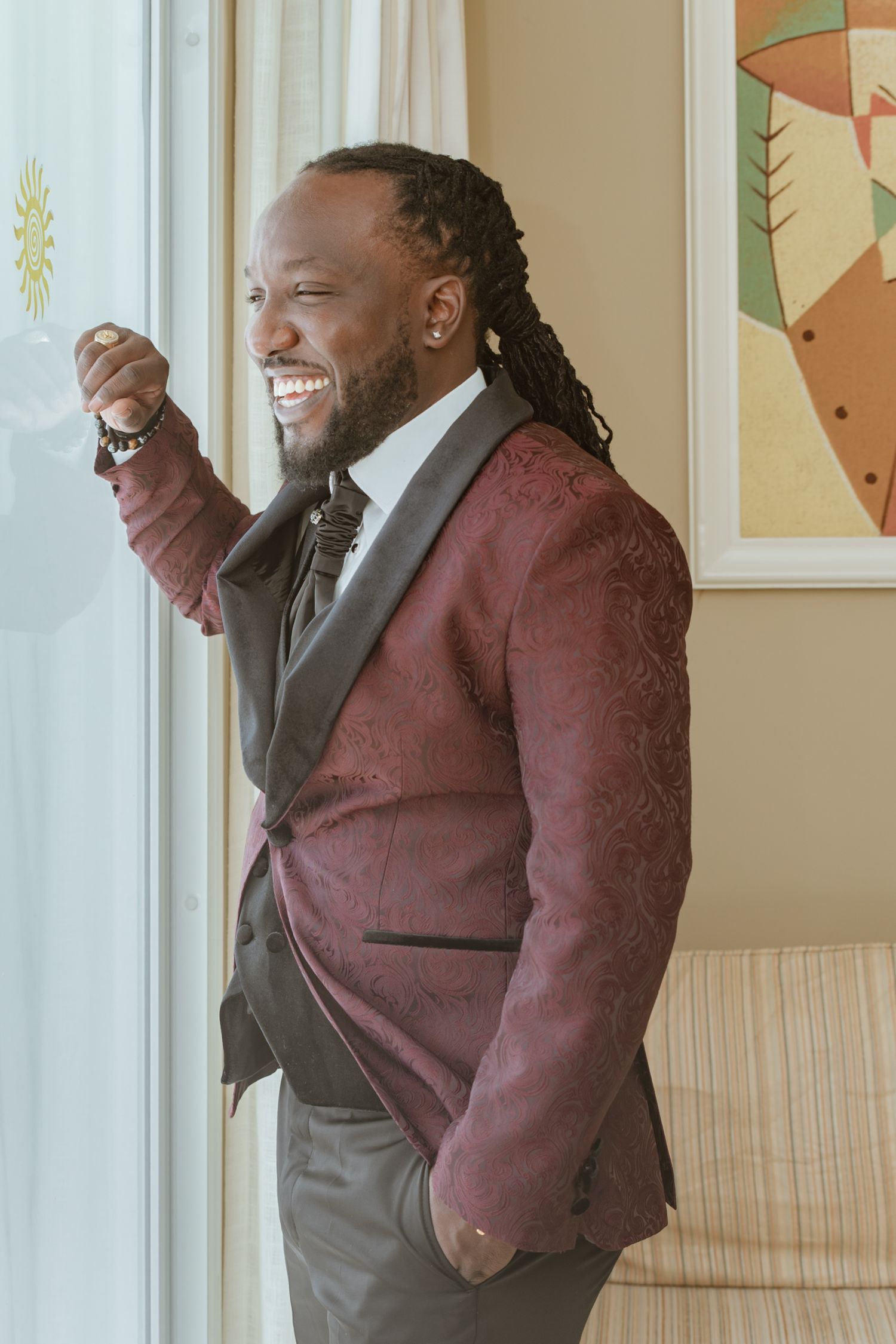 Smiling groom in burgundy floral tuxedo jacket with black lapels laughing by a bright window.
