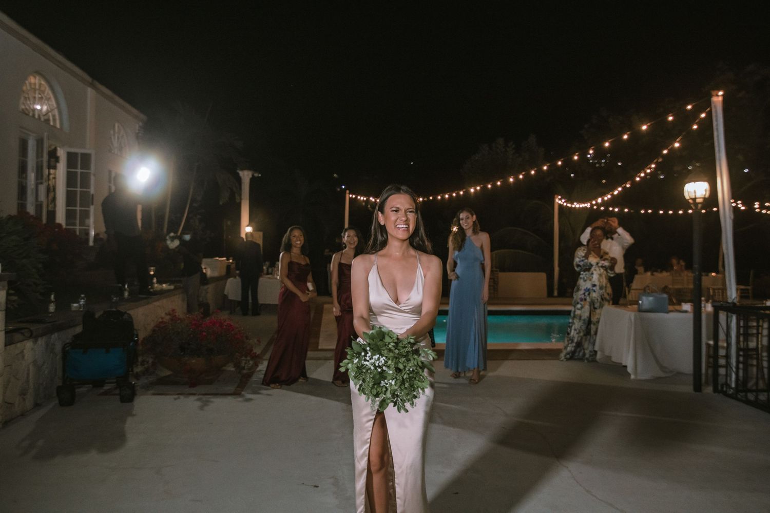 Bride in elegant white gown holding greenery bouquet preparing for bouquet toss at outdoor nighttime reception.