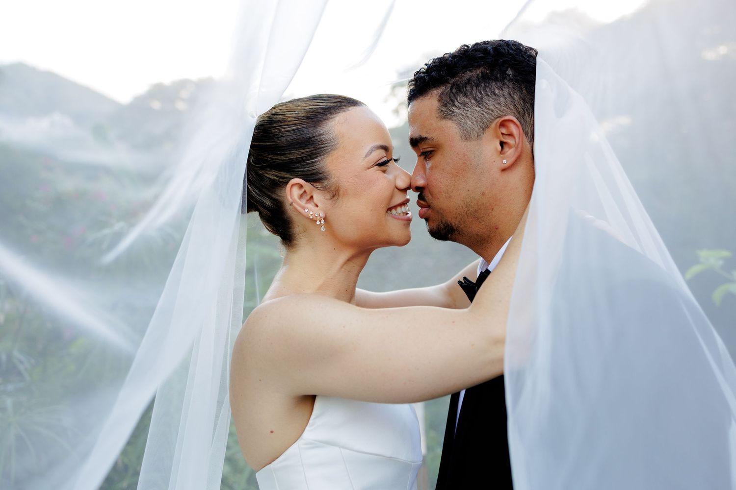 Bride and groom share an intimate nose-to-nose moment beneath a flowing white veil at their wedding.
