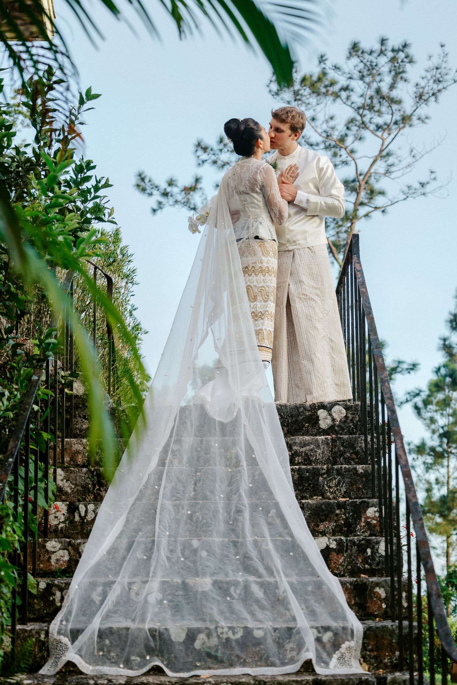 Bride and groom in white attire share a romantic moment on stone steps surrounded by lush greenery.