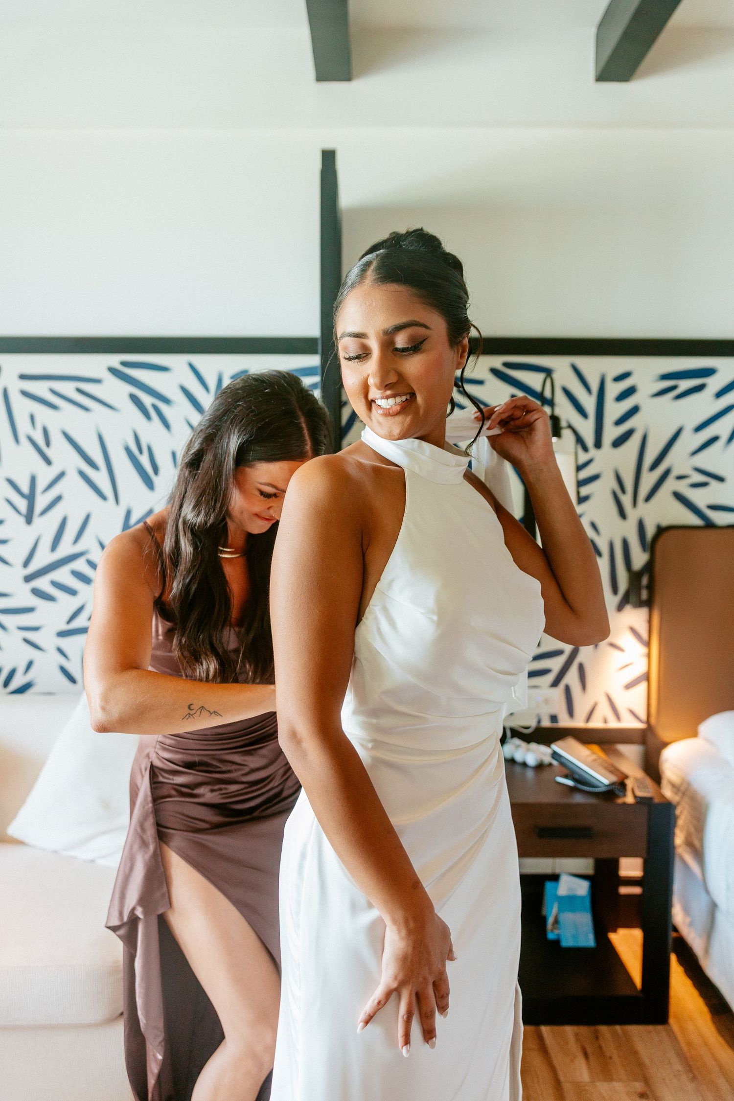 Bridesmaid helping bride into elegant white halter wedding gown in a decorated hotel room.