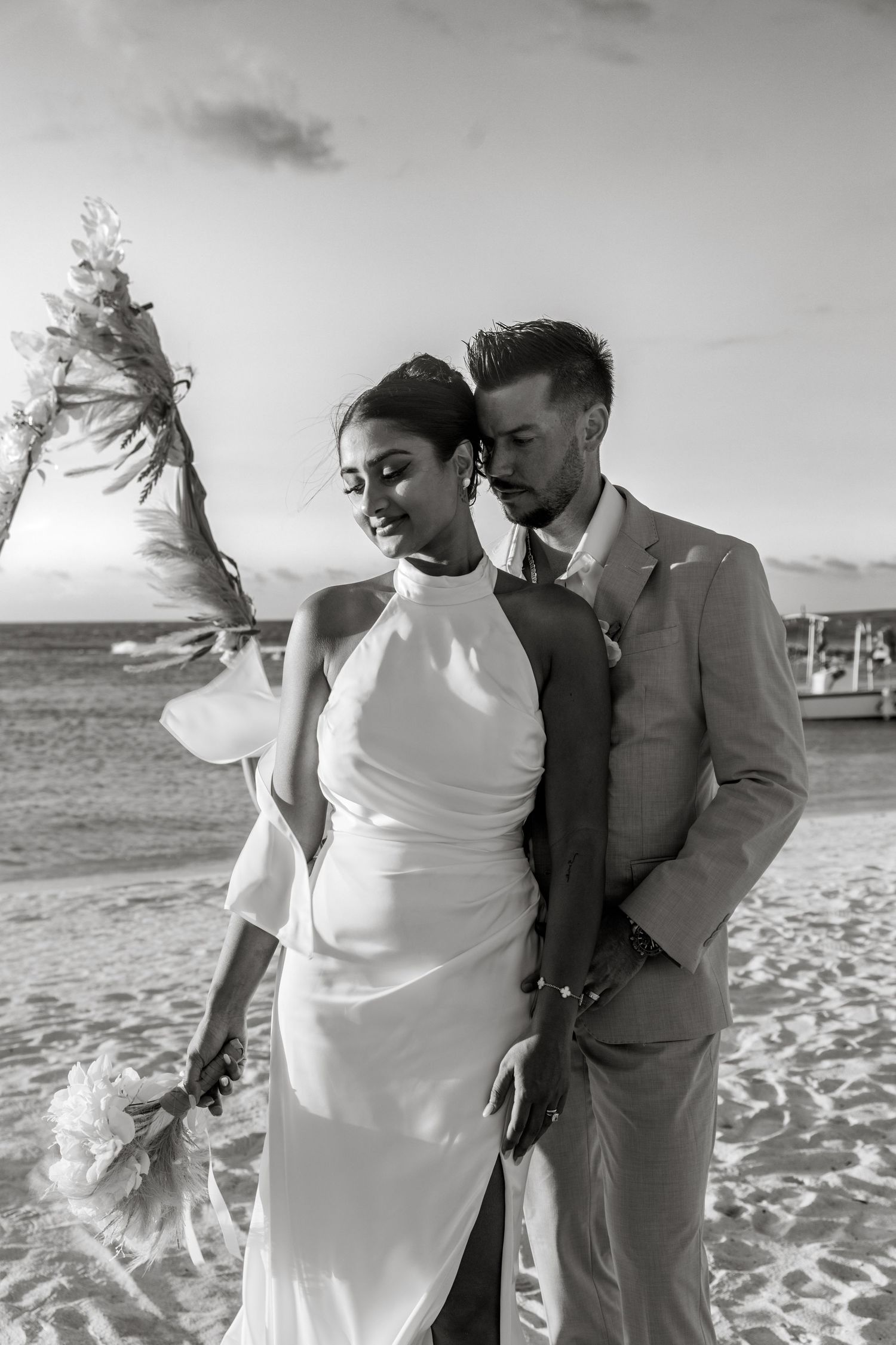 Black and white photo of bride and groom embracing on a windy beach near a decorated wedding arch at dusk.