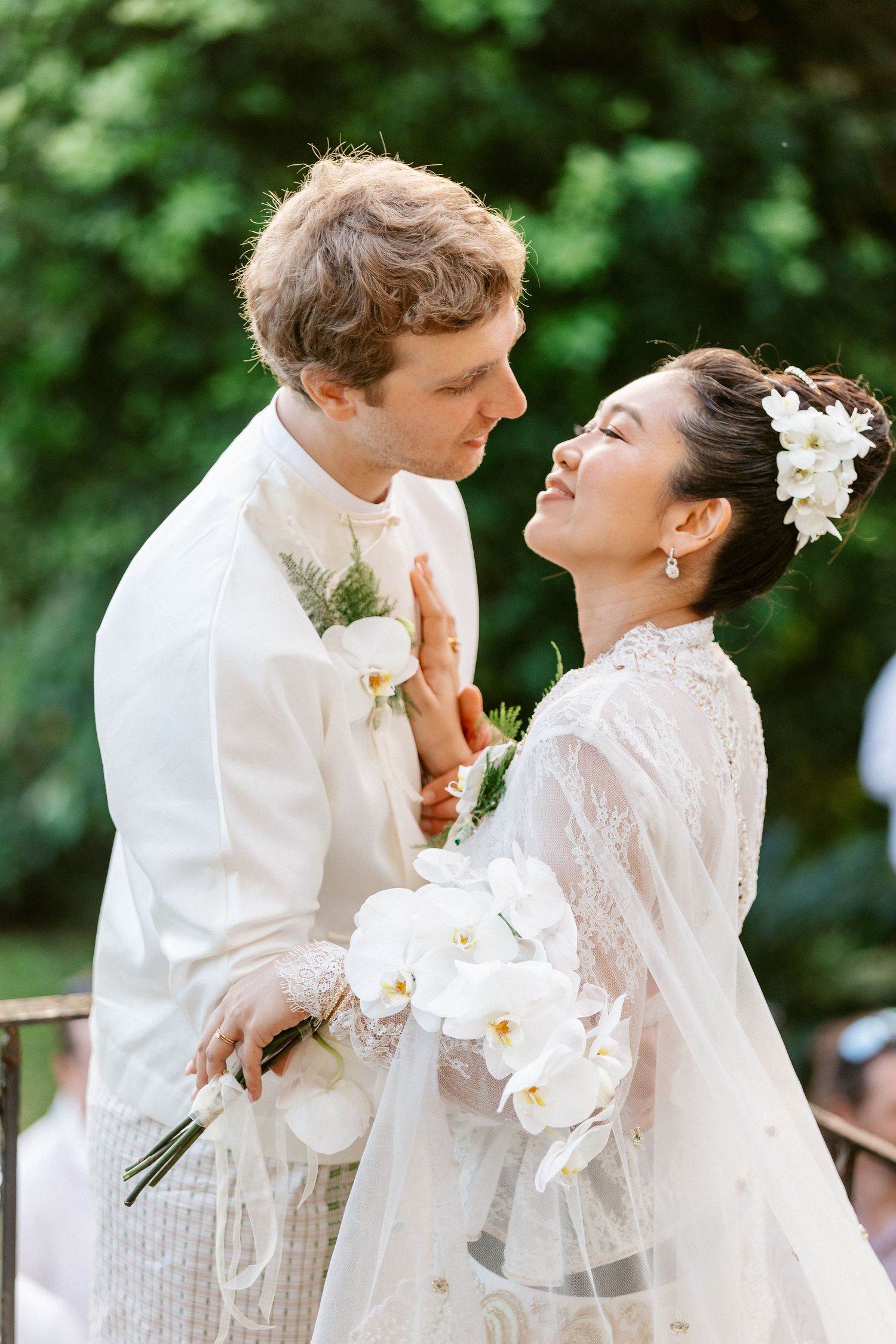 Bride and groom in white wedding attire sharing a tender moment outdoors with orchid bouquet.