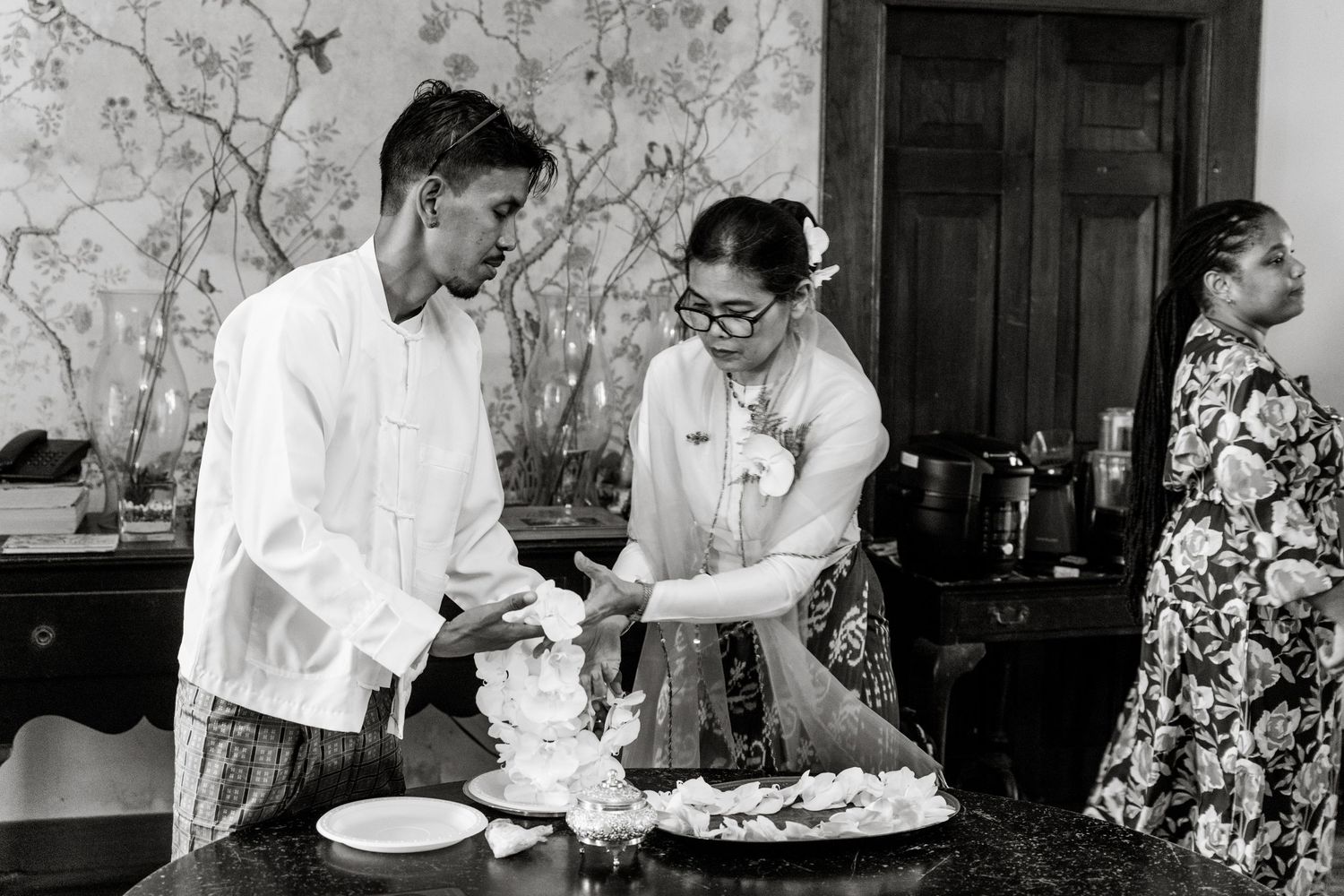 Black and white photo of a couple preparing food together in a kitchen, with a woman in glasses and floral dress.