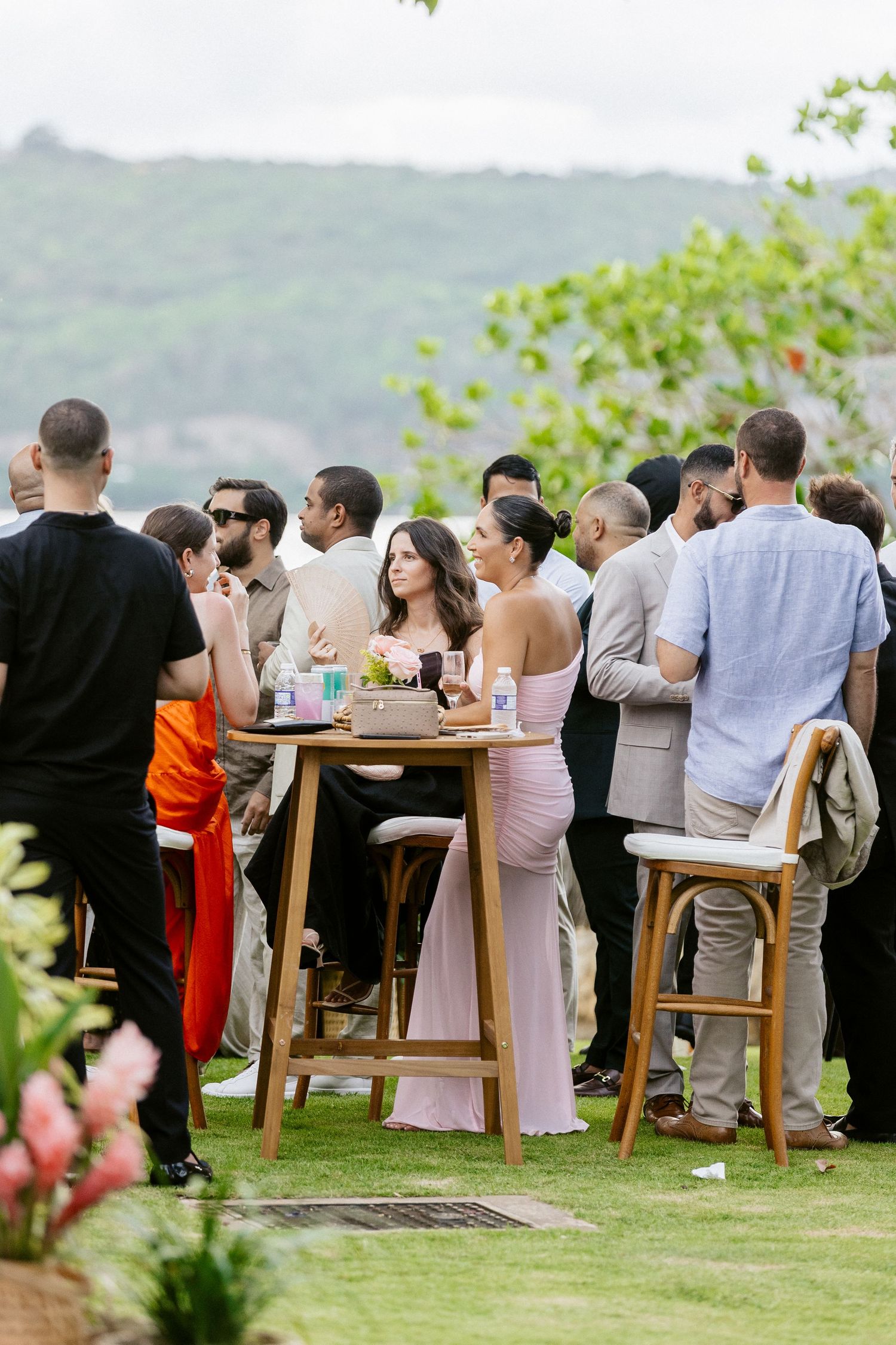 Guests mingle at an outdoor wedding cocktail hour with lush green hills in the background.