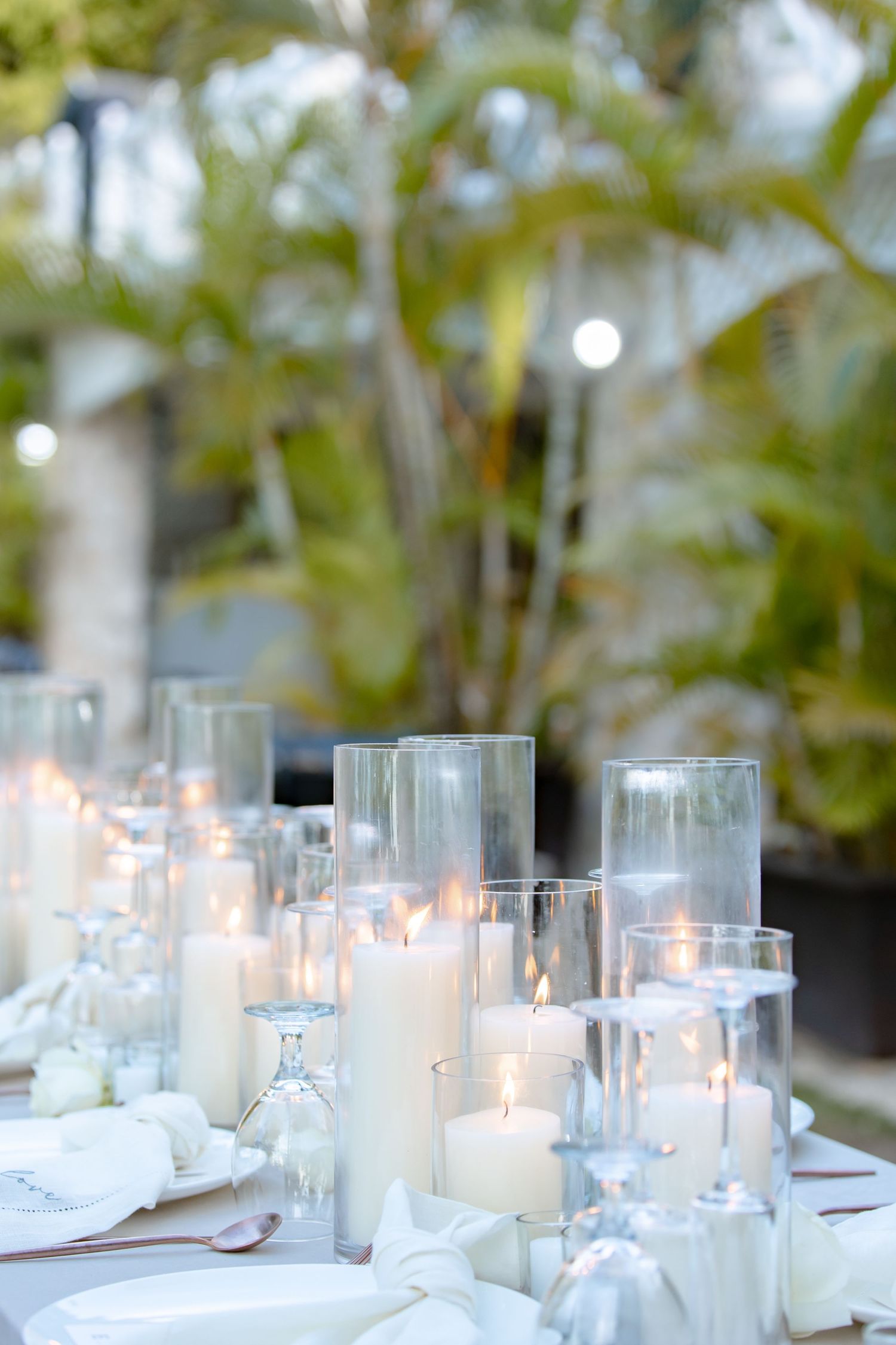 Glowing white pillar candles in glass hurricane vases decorating an elegant outdoor wedding reception dinner table.