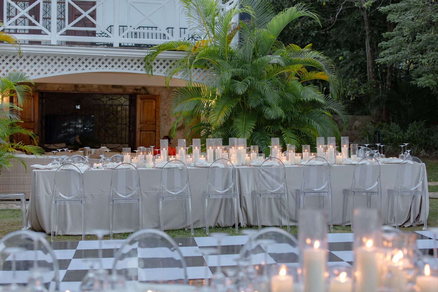 Elegant outdoor wedding reception with long white draped table, candles, and lush tropical greenery at dusk.