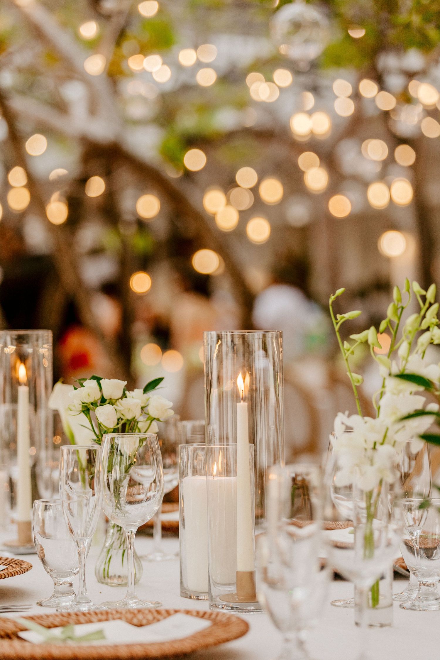 Enchanting outdoor wedding reception table with string lights bokeh, white roses, pillar candles, and wicker charger plates.