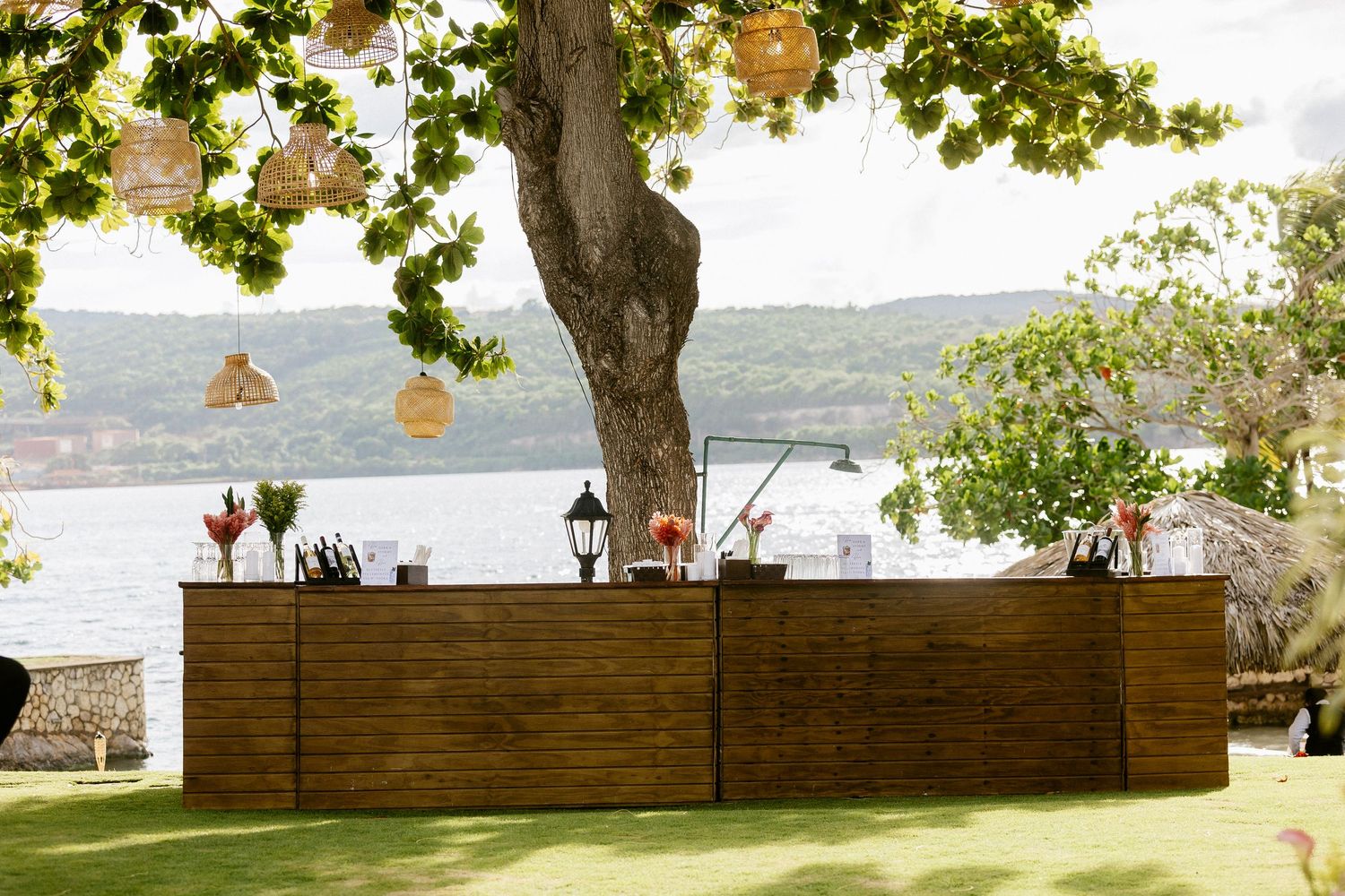 Rustic wooden outdoor bar setup under a tree with hanging lanterns and scenic waterfront views at a wedding reception.