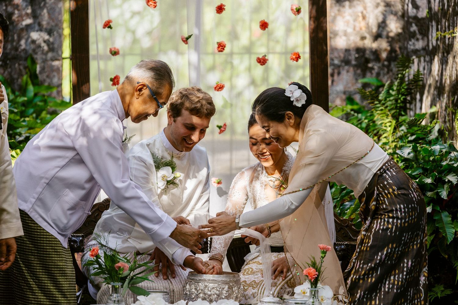 Family members joyfully participate in traditional Burmese wedding ceremony outdoors surrounded by lush greenery.