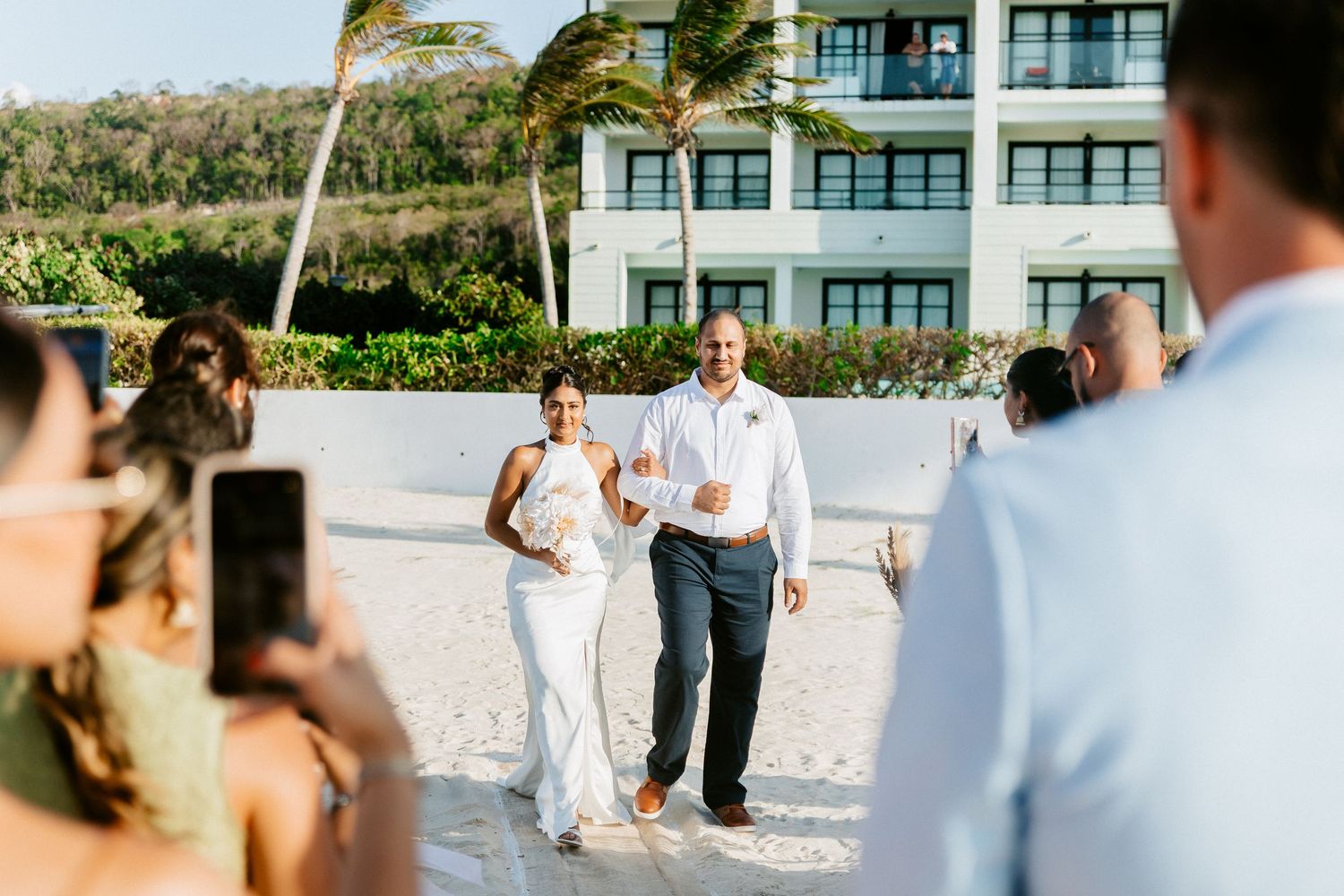 Couple walks joyfully on sandy beach after wedding ceremony with tropical resort and palm trees in background.