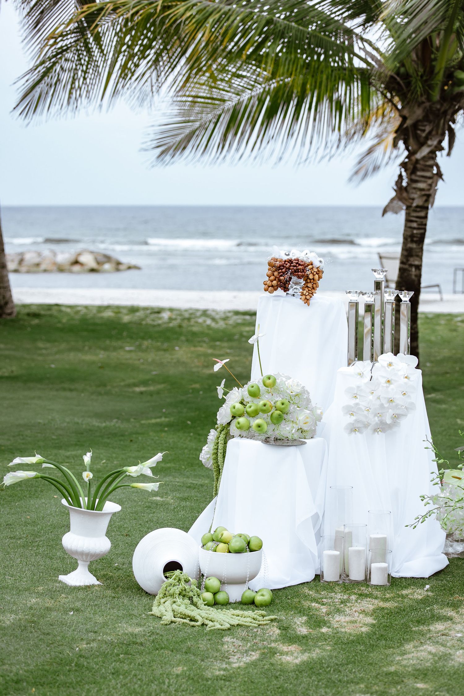 Elegant beachside wedding setup with white draping, green apples, calla lilies, and candles on a tropical lawn.