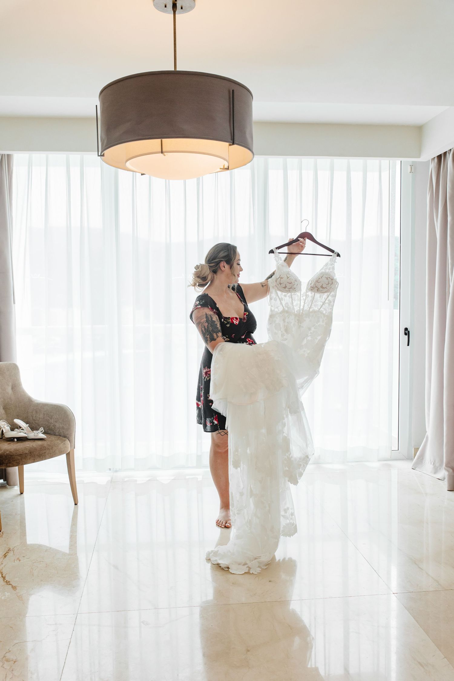 Bride holding her lace wedding dress on a hanger in a bright hotel room before the ceremony.