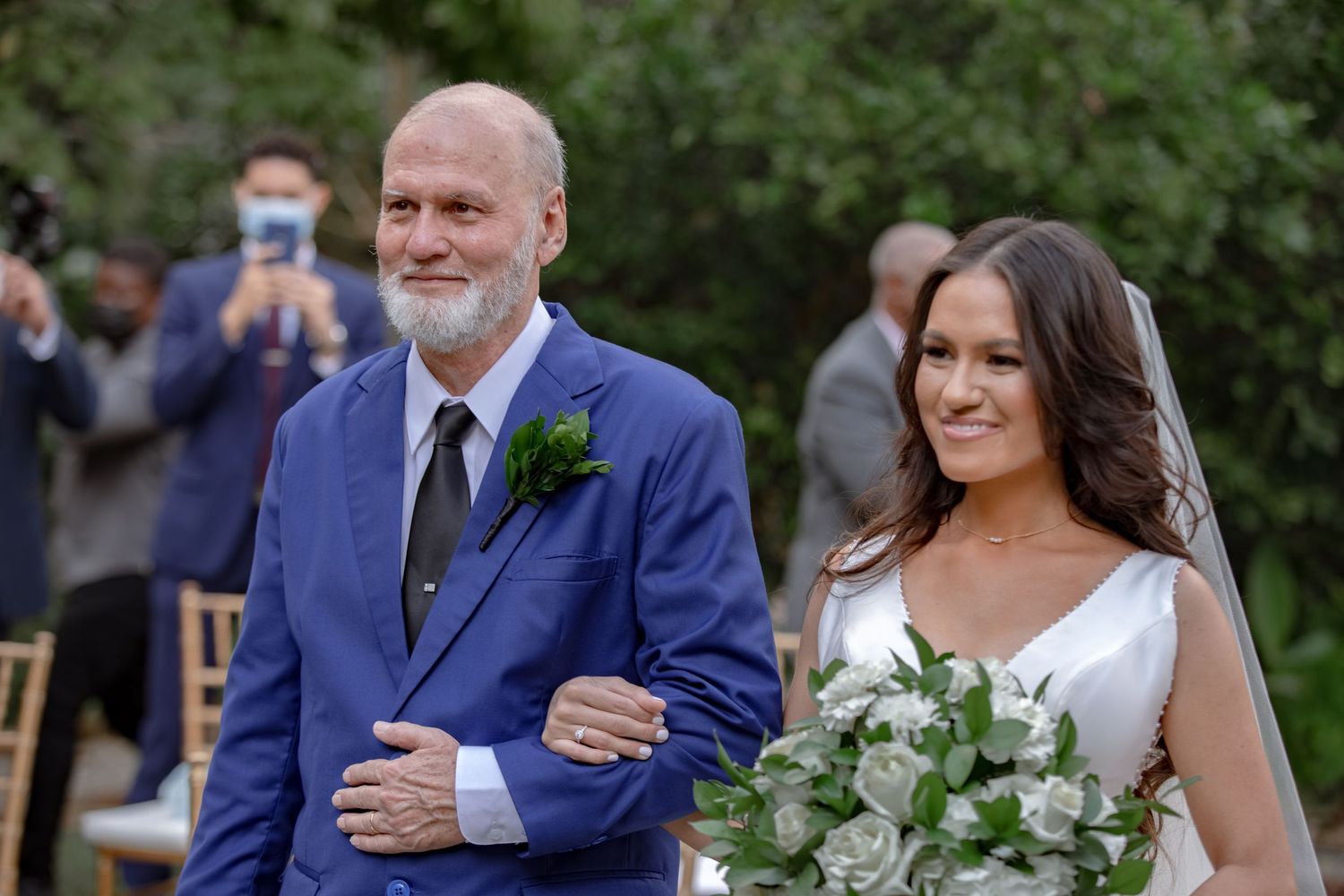 Father walking bride down the aisle at outdoor wedding ceremony, bride holding bouquet.