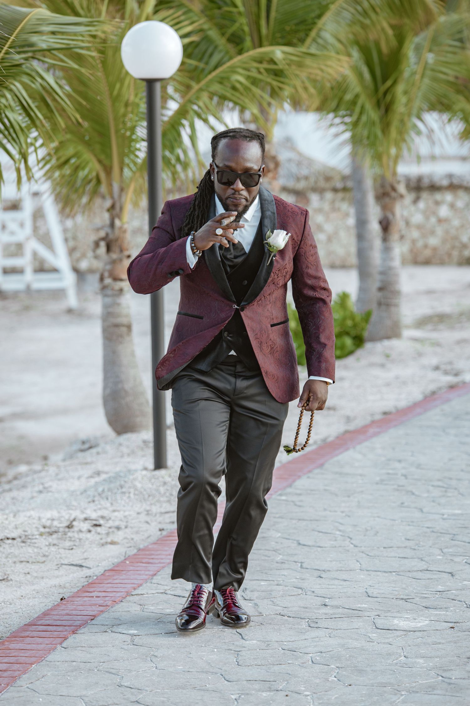 Stylish groom in burgundy tuxedo jacket poses confidently on tropical pathway holding pocket watch.