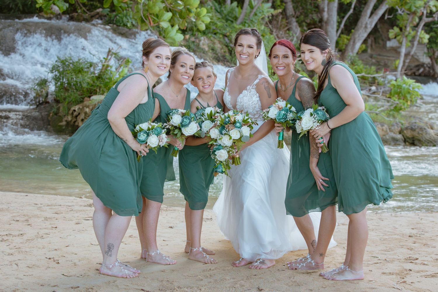 Bride with four bridesmaids in green dresses holding white bouquets on a tropical beach.