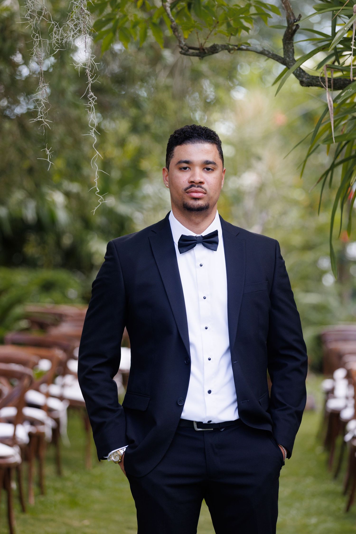 A dapper groom in a black tuxedo with bow tie stands confidently at an outdoor wedding ceremony aisle.