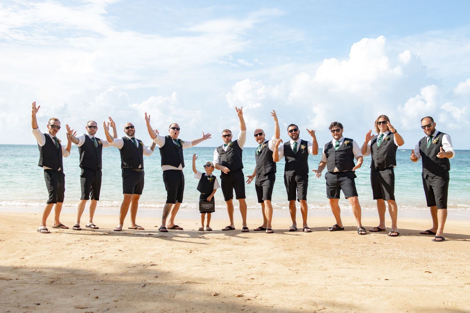 Groomsmen in black vests and shorts celebrate on a tropical beach, raising hands and cheering together.