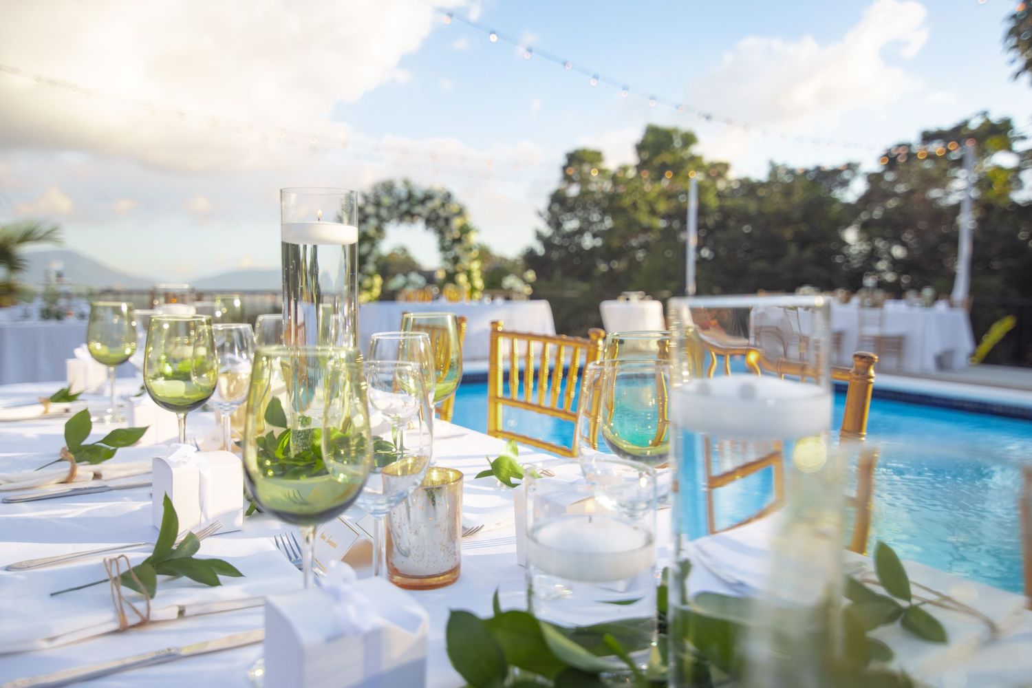 Outdoor wedding reception table setting beside pool with green floral decor and gold chairs.