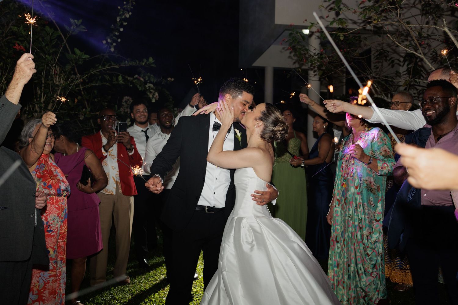 Newlyweds kiss passionately as wedding guests celebrate with sparklers during an outdoor evening reception.