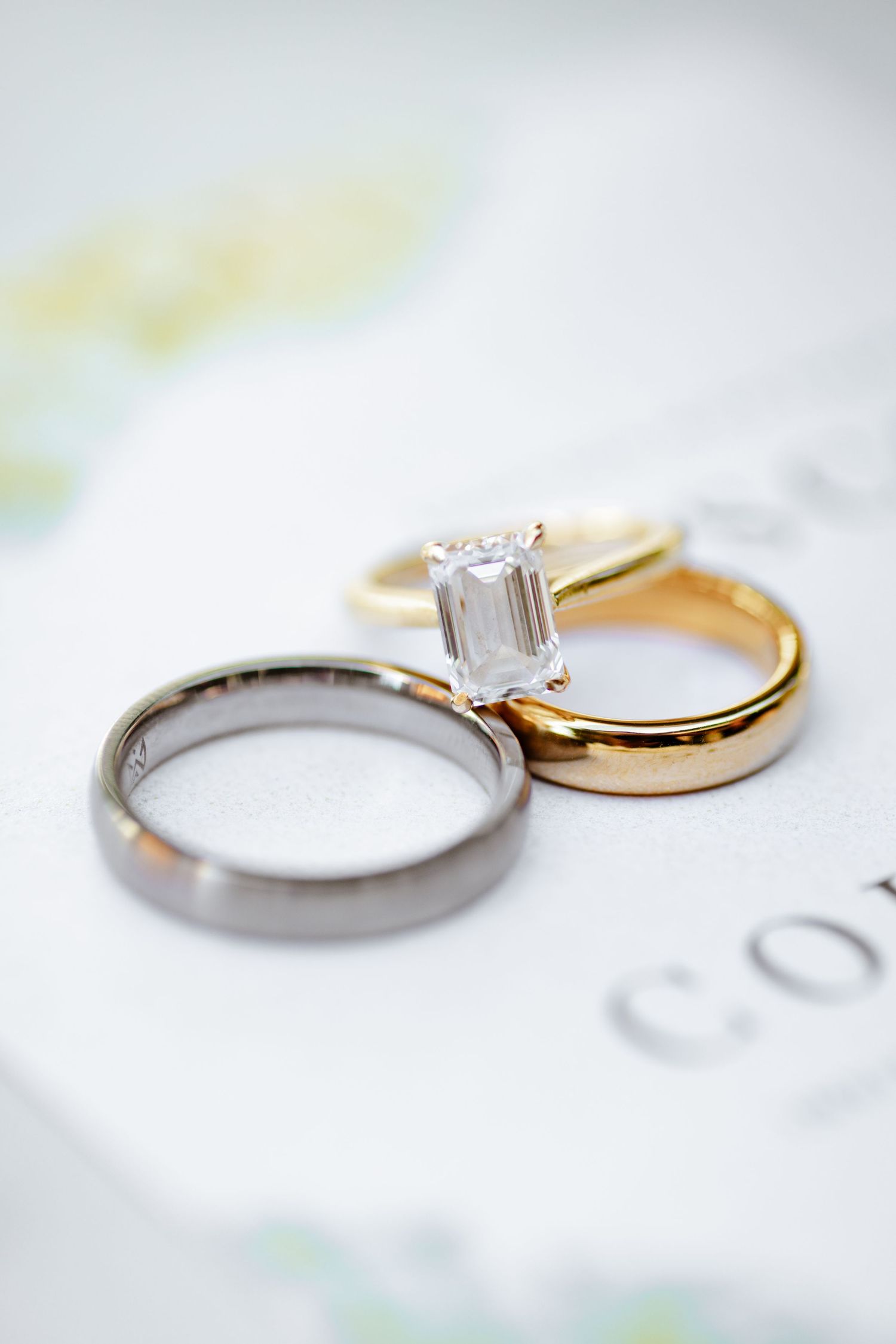 Close-up of emerald cut diamond engagement ring with gold band and silver wedding band on floral invitation.