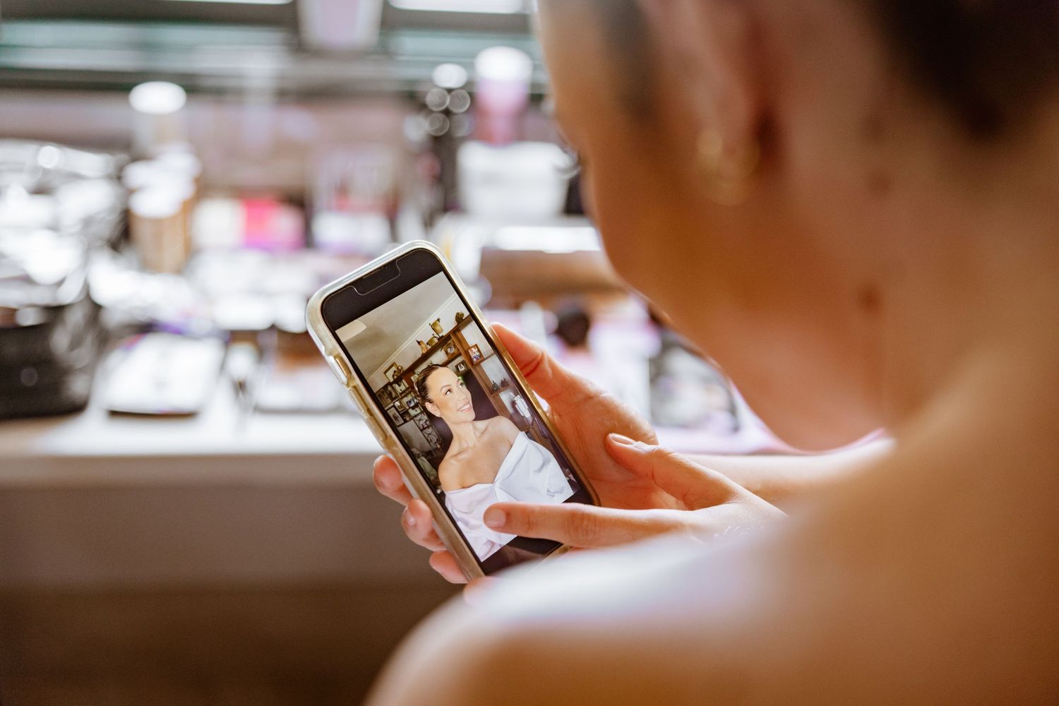Bride viewing wedding photo on smartphone at makeup vanity during bridal preparations.