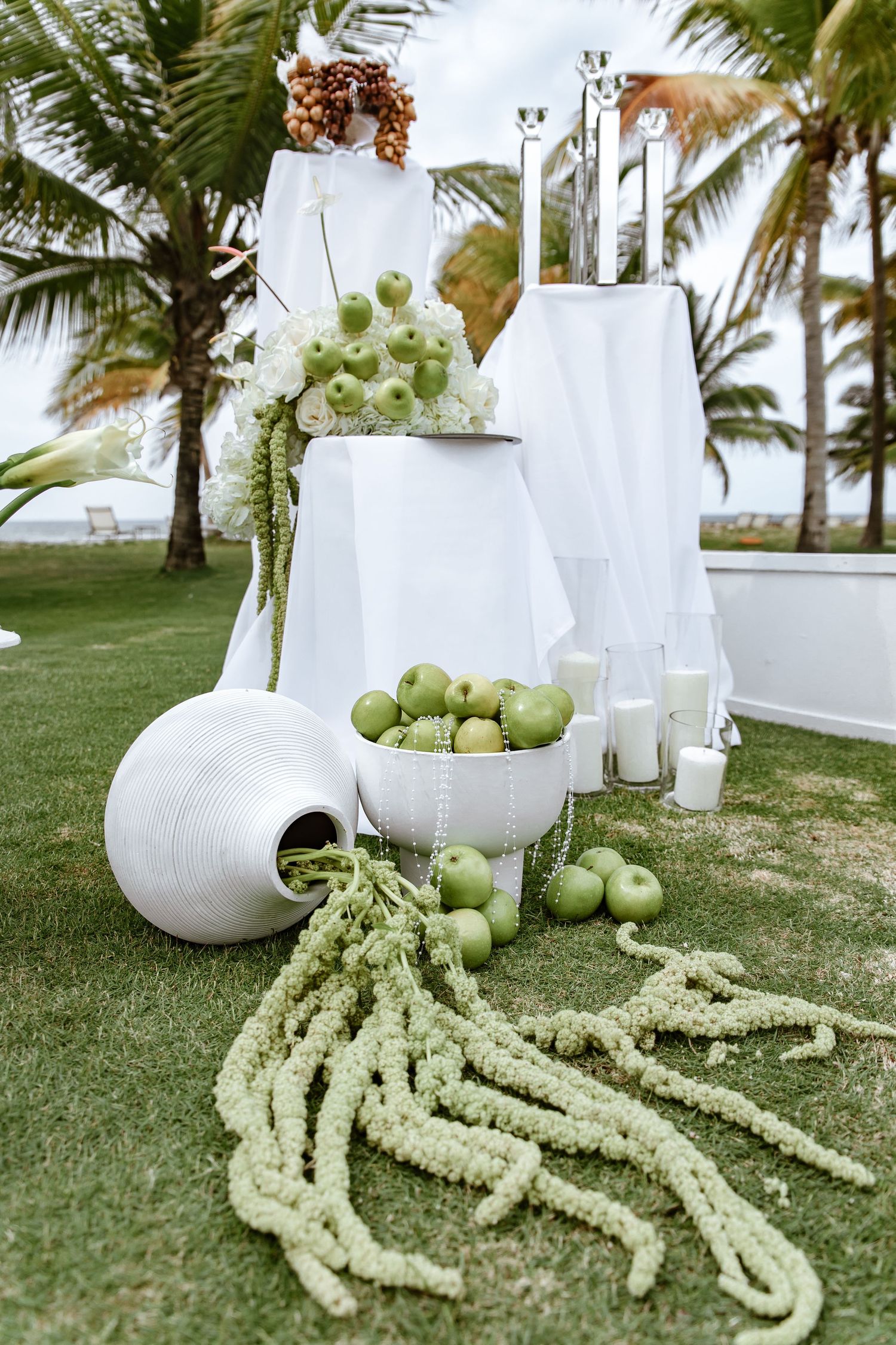 Elegant outdoor wedding decor with green apples, white flowers, candles, and palm trees in a tropical setting.