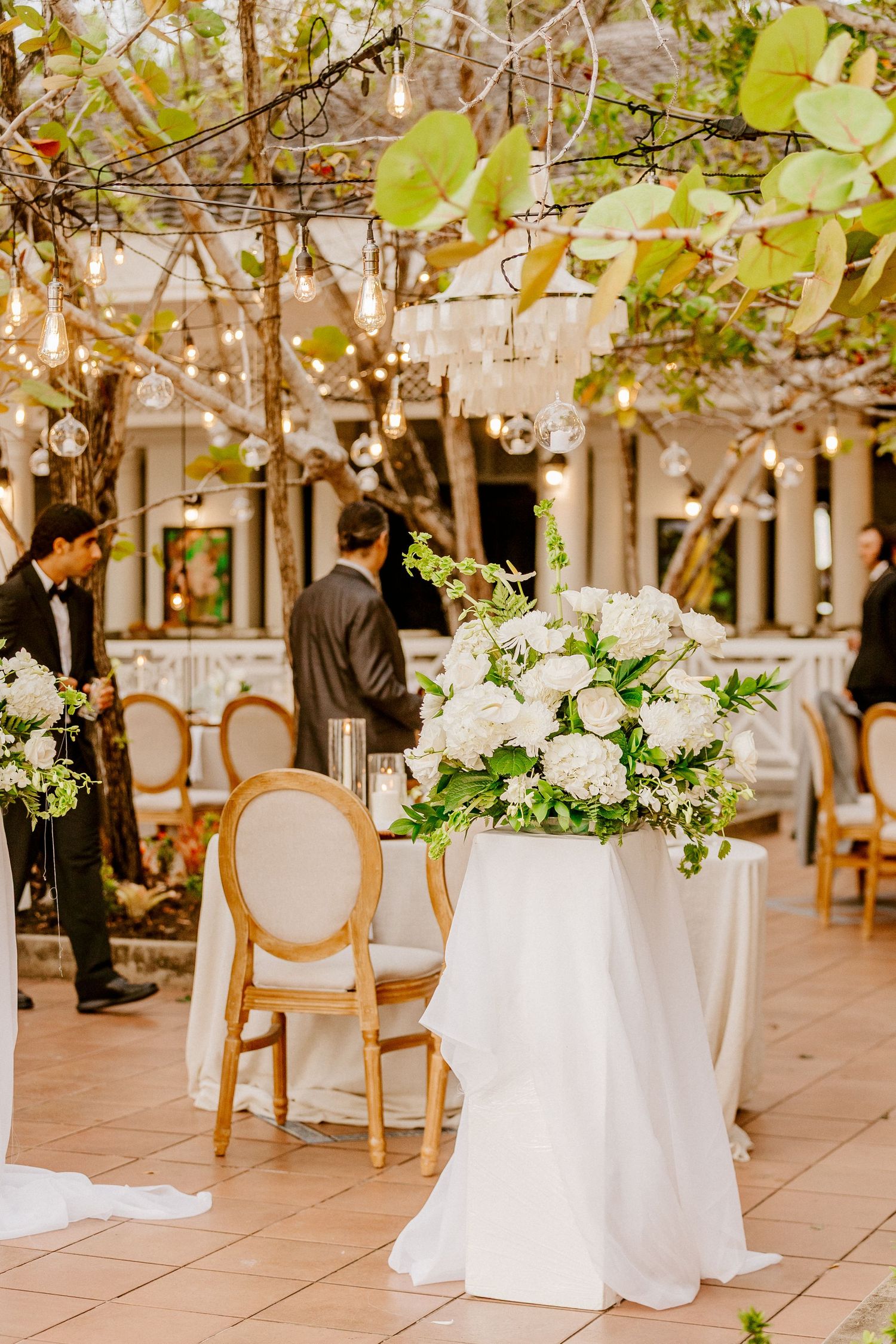 Elegant wedding reception decor with white floral centerpiece, gold chairs, and string lights under tree canopy.