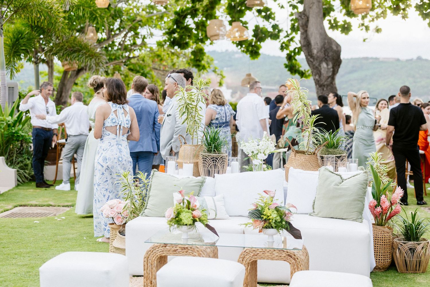 Elegant outdoor wedding reception with white lounge furniture, tropical plants, and guests mingling on a lush green lawn.