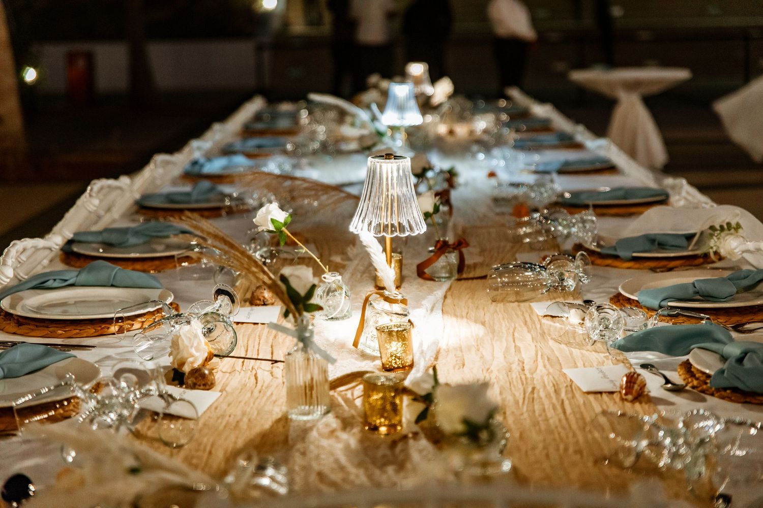 Elegant wedding reception table set with gold chargers, blue napkins, and glowing candles.