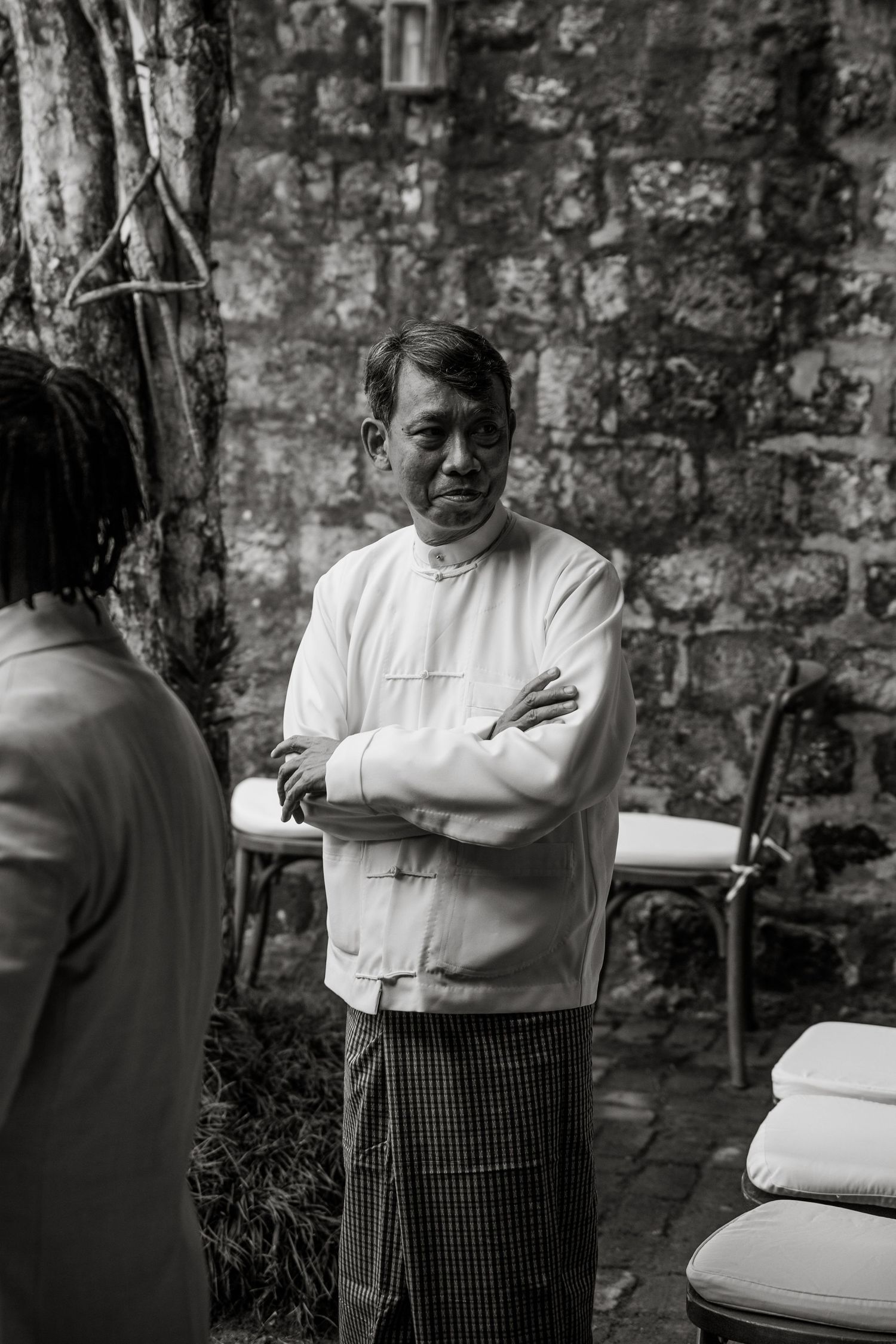 Black and white photo of a man in traditional attire with arms crossed, standing outdoors near a stone wall.