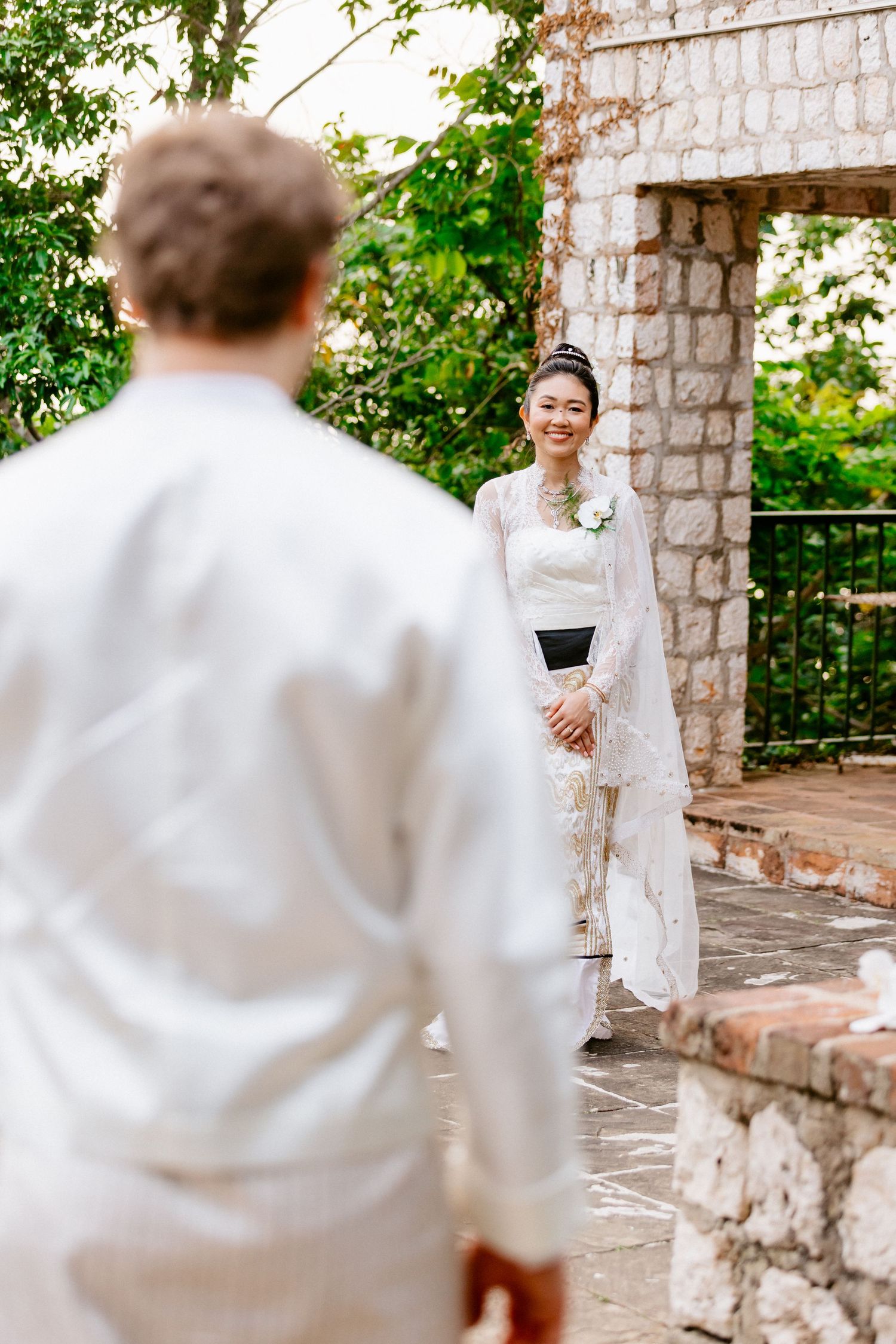 Bride in traditional white lace Asian dress smiles at groom during outdoor stone venue first look.