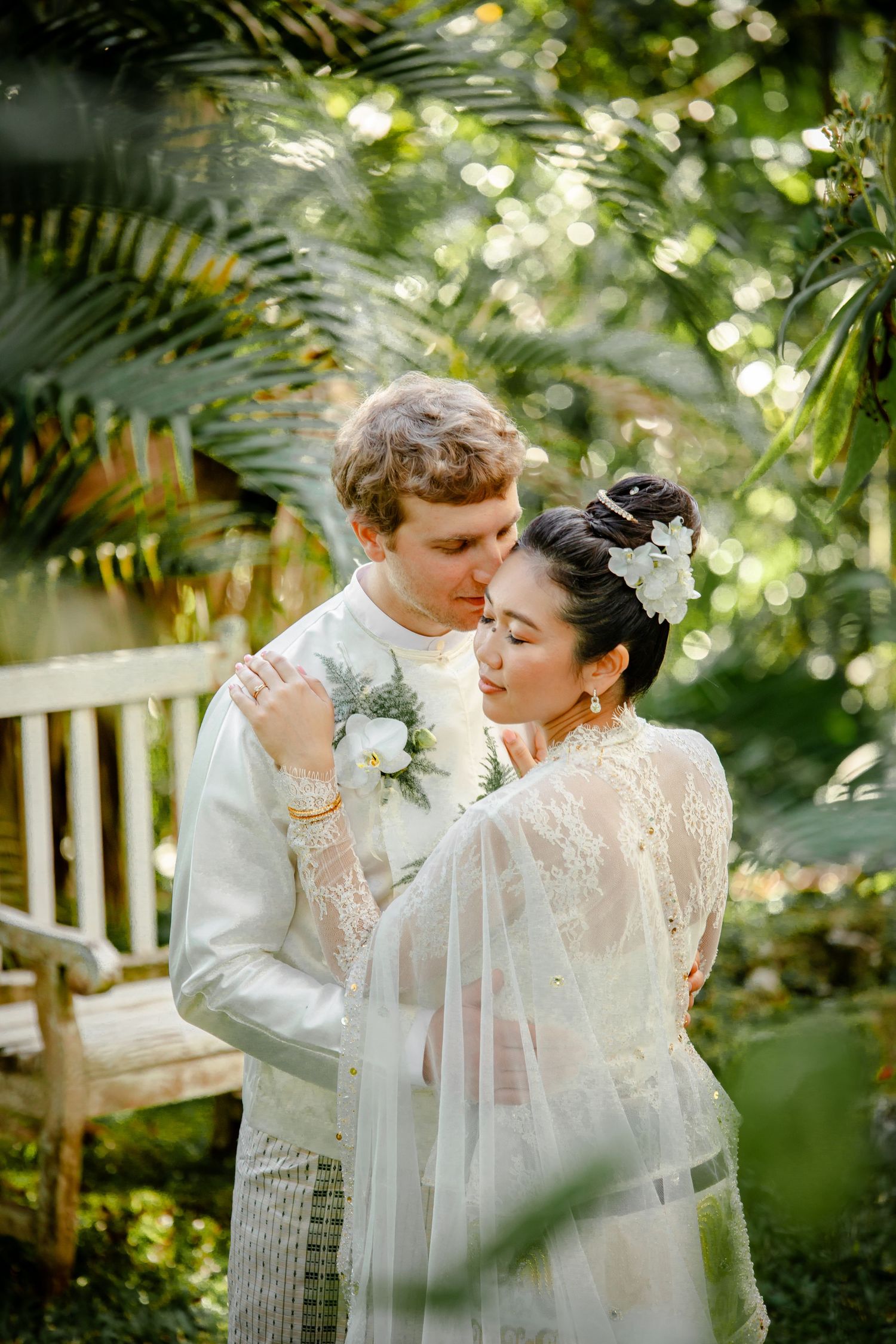 Bride and groom embracing in lush tropical garden, bride wearing lace gown with floral headpiece.